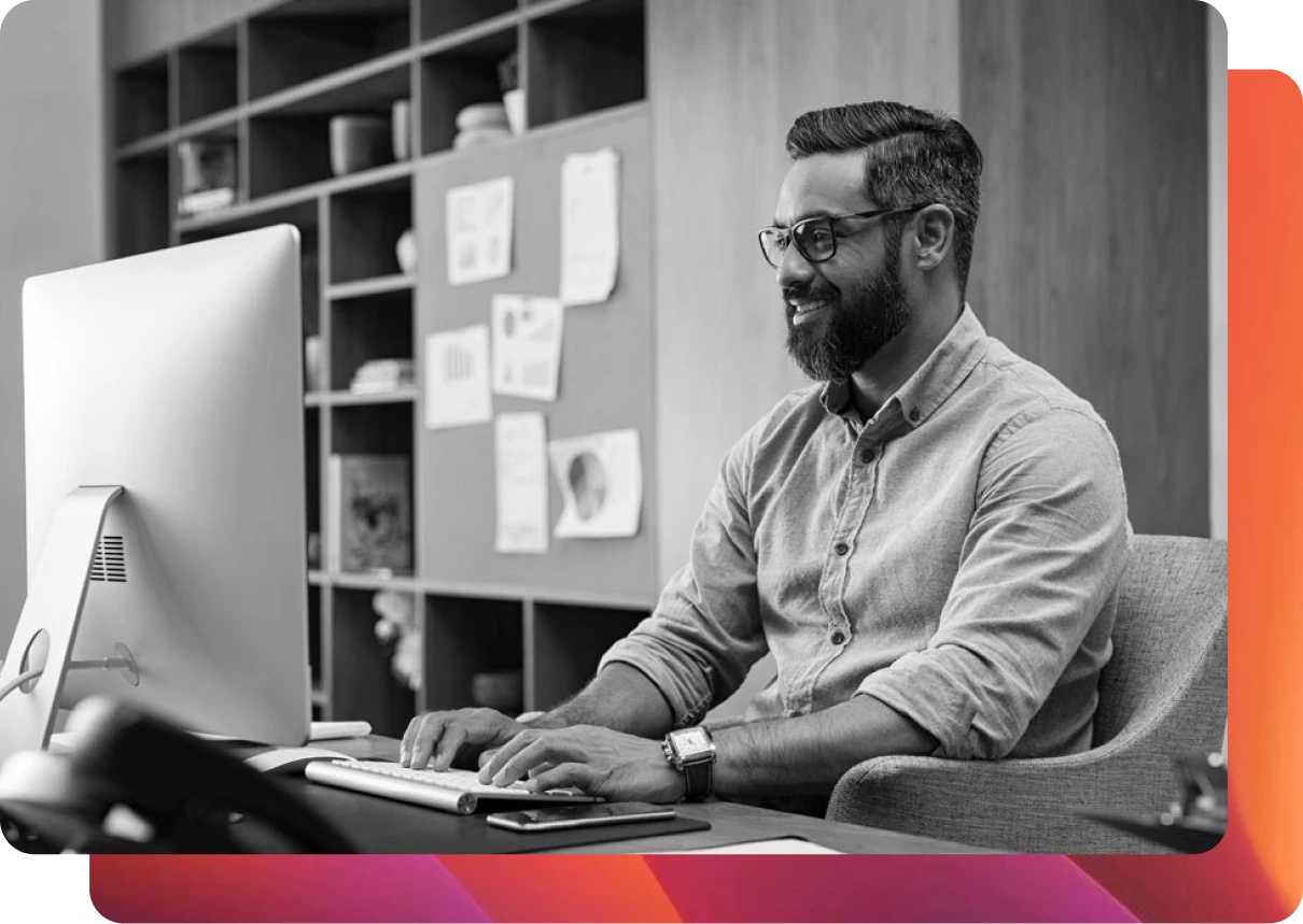 Bearded man with glasses sitting down at a desk working on an iMac in black and white