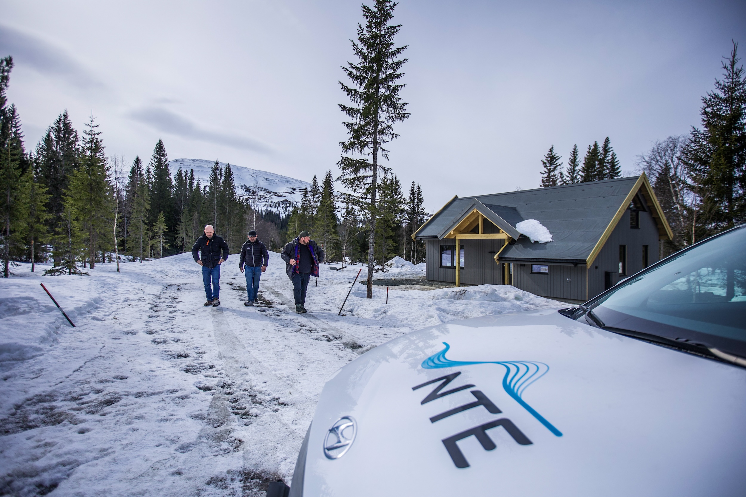 Tre menn går mot en bil med NTE-logoen på panseret. I bakgrunnen en hytte, snø, fjell og skog. 