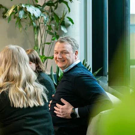 Geir Arne Rø Bilde av en mann som sitter i en sofa i et kontorlandskap og ser mot kamera og smiler. Ved siden av ser man ryggen til to kvinnelige kollega.
