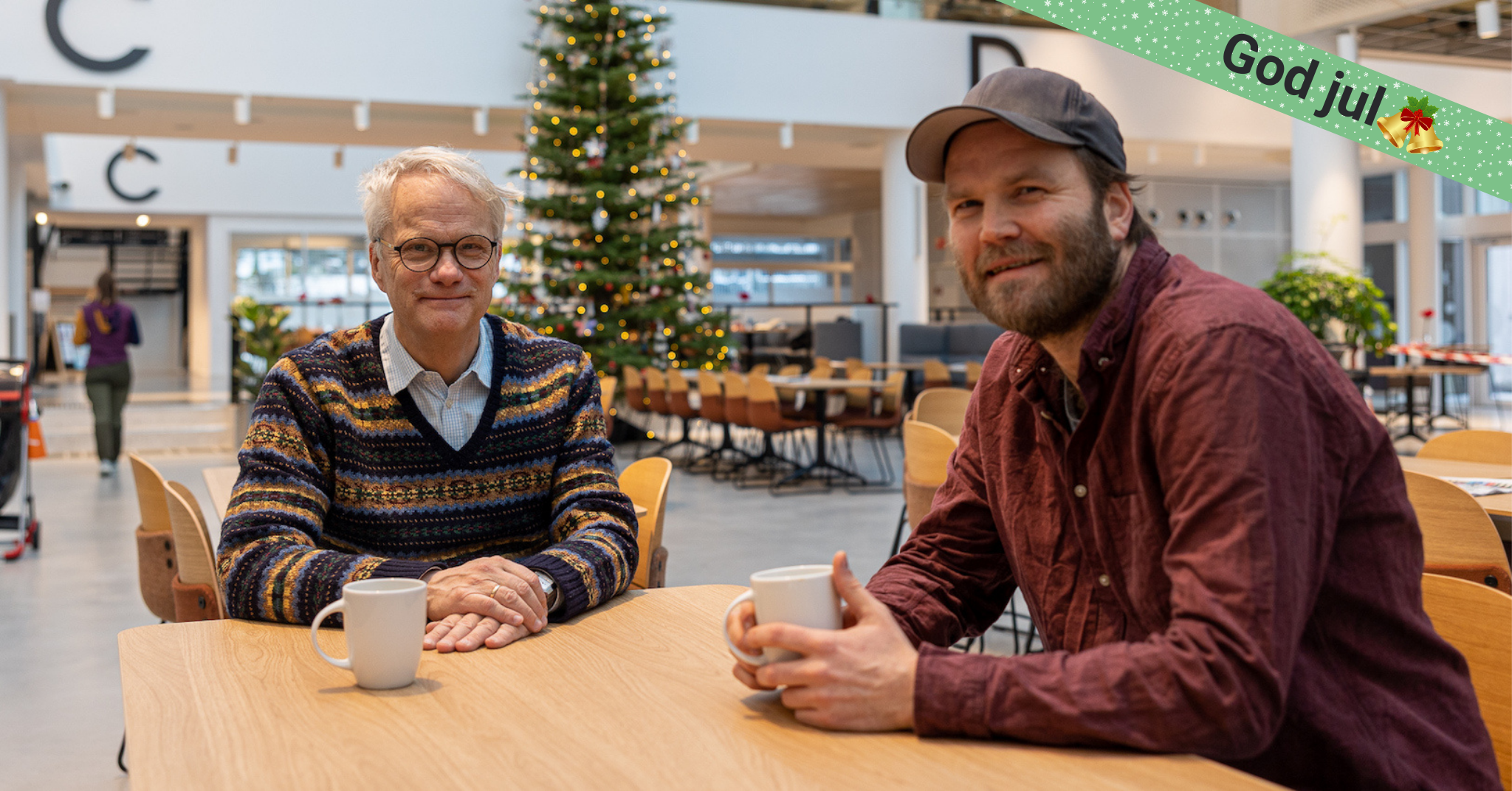 To menn med hver sin kaffekopp smiler, med et stort juletre i bakgrunnen