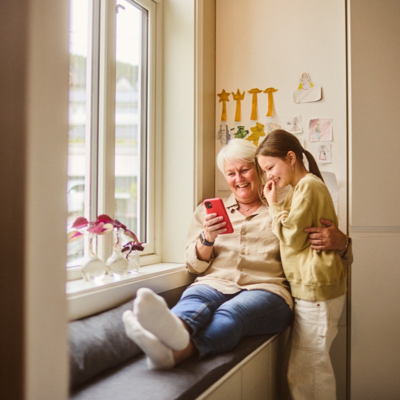 Bilde av en bestemor som sitter på en benk ved et vindu og smiler mens hun holder rundt et barn. De smiler og ser på mobilen sammen.