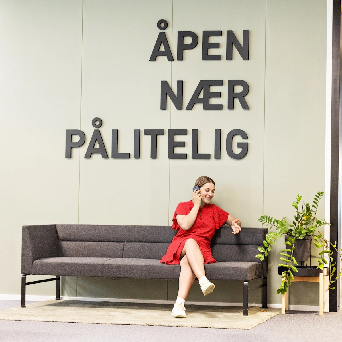 En dame i kjole sitter i en sofa og snakker i telefonen. Over henne står ordene Åpen nær pålitelig En dame i kjole sitter i en sofa og snakker i telefonen. Over henne står ordene Åpen nær pålitelig