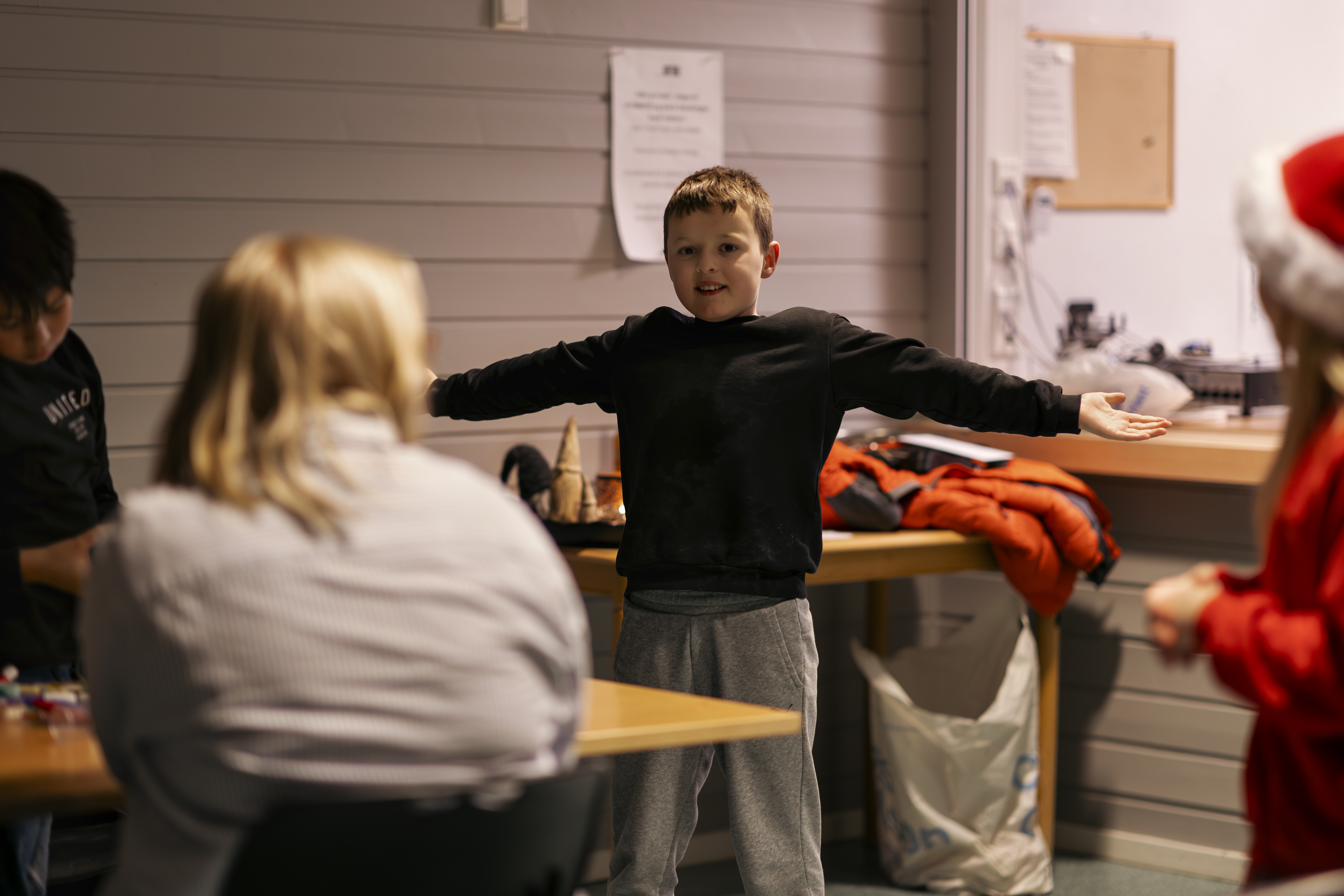 En smilende gutt slår ut med armene. Vi ser ryggen på to personer og en jente med nisselue. 