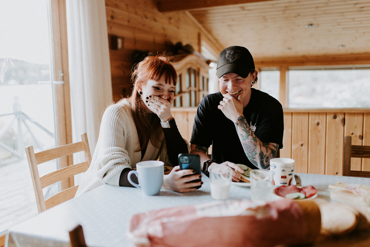 Bilde av en mann og en dame som sitter ved et bord. Damen holder en mobiltelefon i hånda og begge ser på skjermen og smiler.