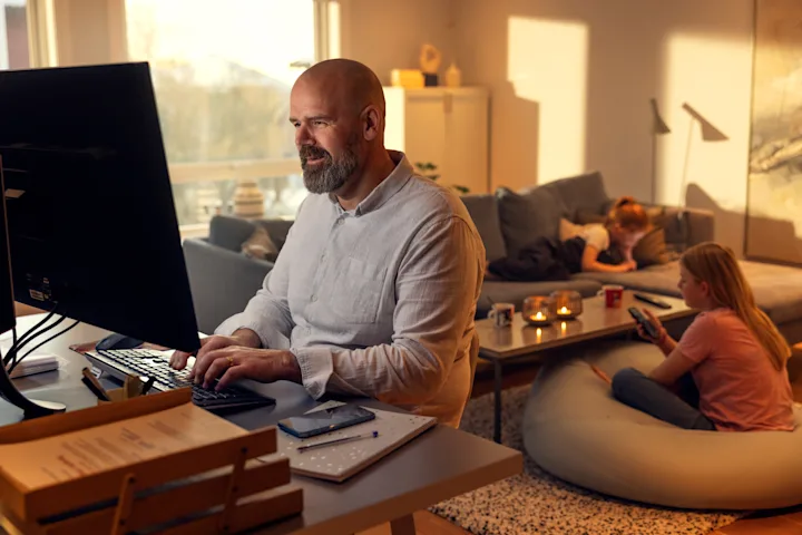 Mann sitter ved datamaskinen på stua med barn i bakgrunnen. Mann sitter ved datamaskinen på stua med barn i bakgrunnen.