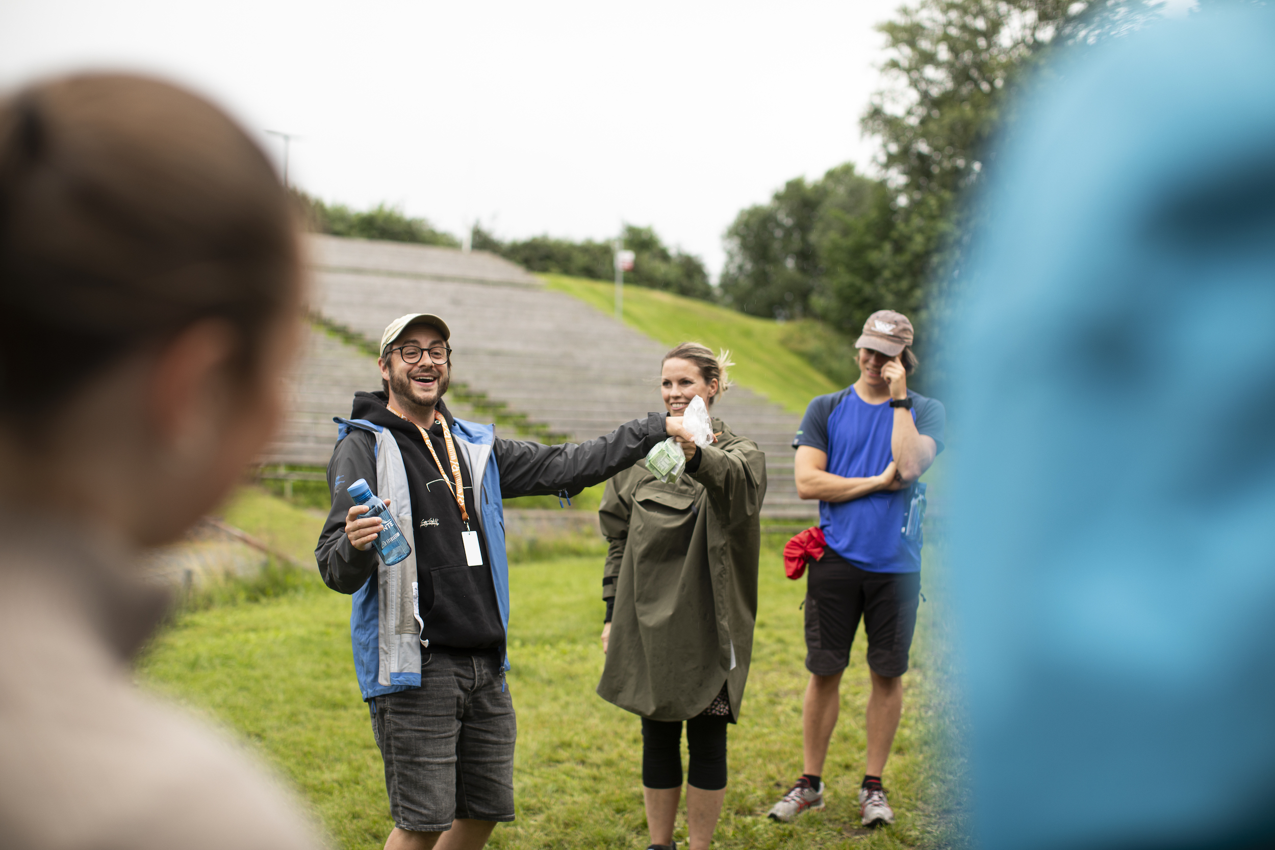 Mann med caps holder opp en vannflaske og en liten pose mens en kvinne og en mann ser på