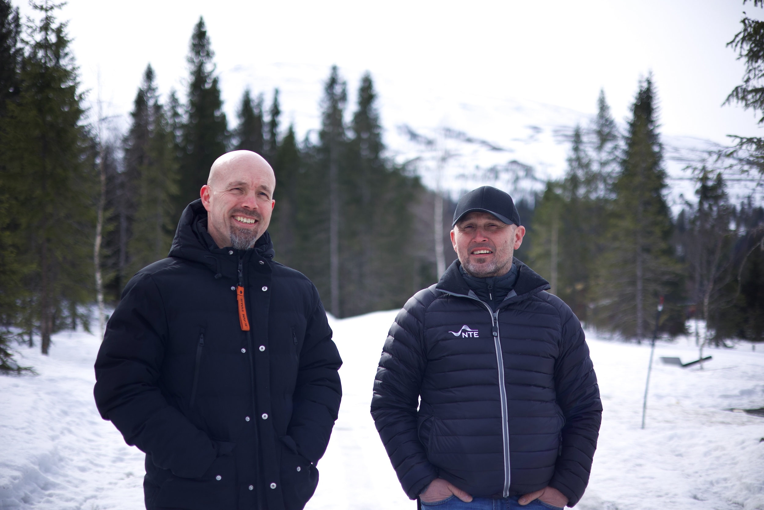 To smilende menn; en med caps, står på en skogsvei i snø med snødekte fjell og grantrær bak seg