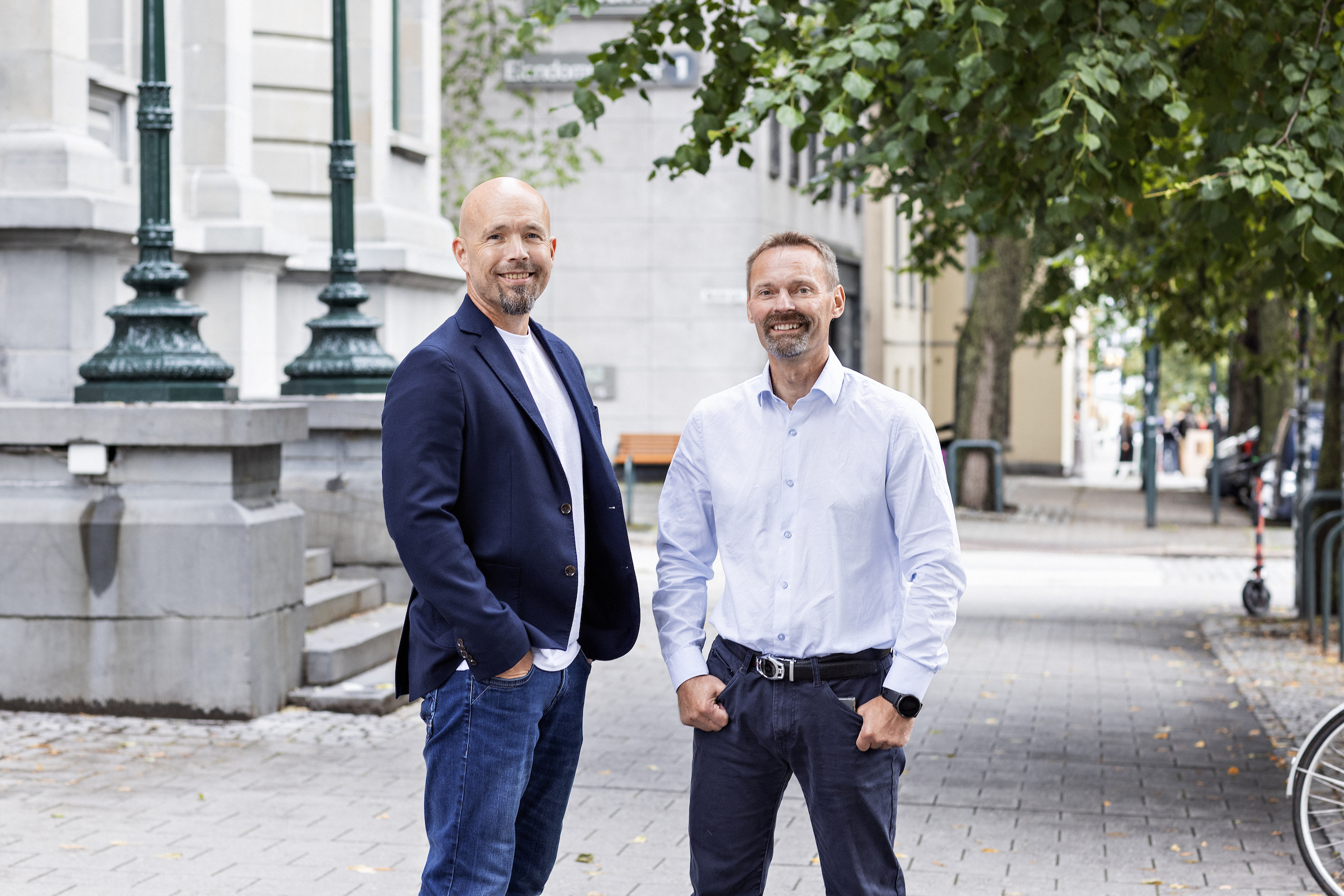 To smilende menn står i bybildet i Trondheim