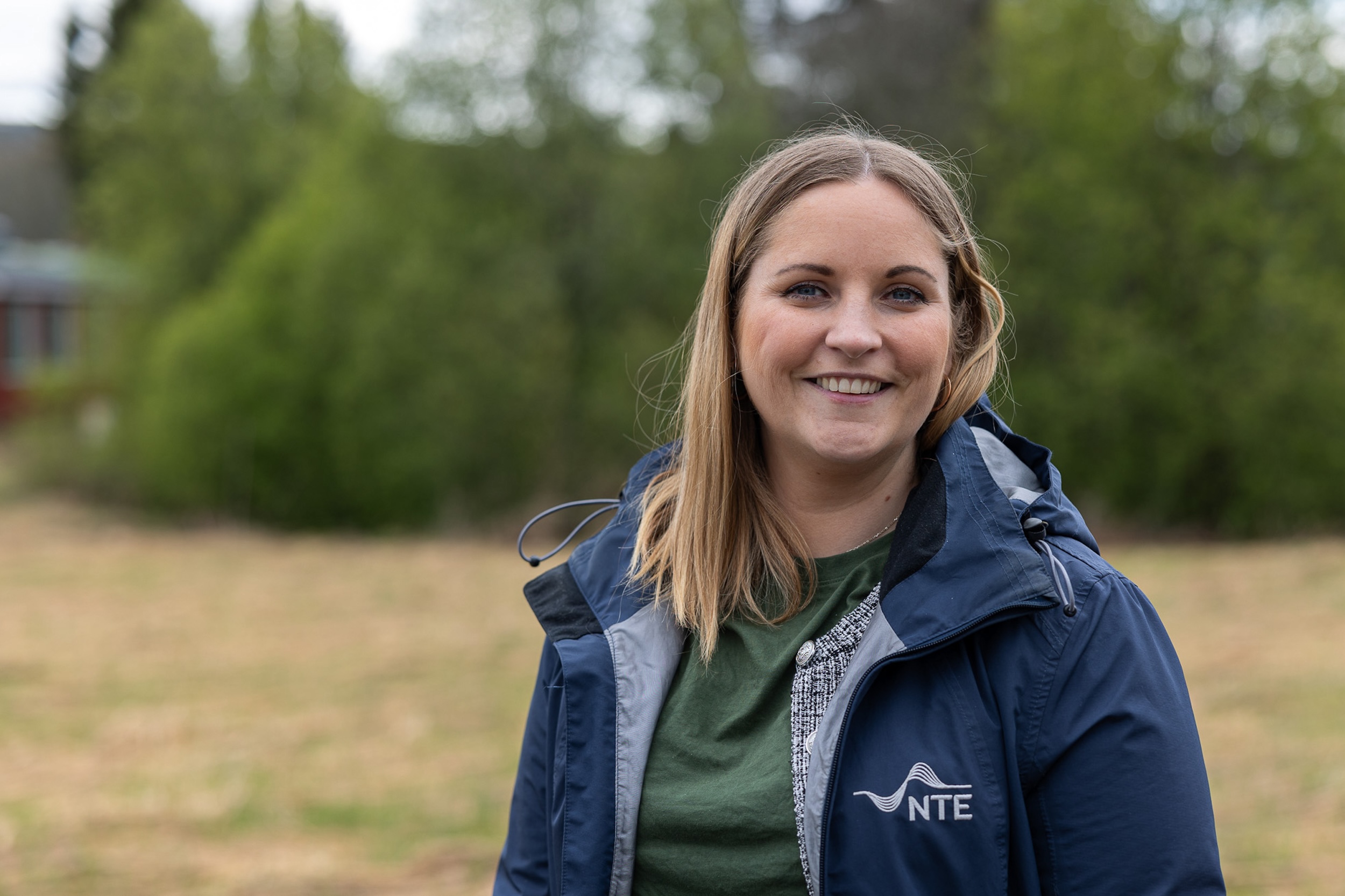 Smilende dame med halvlangt hår og jakke med NTE-logoen på står foran en treklynge