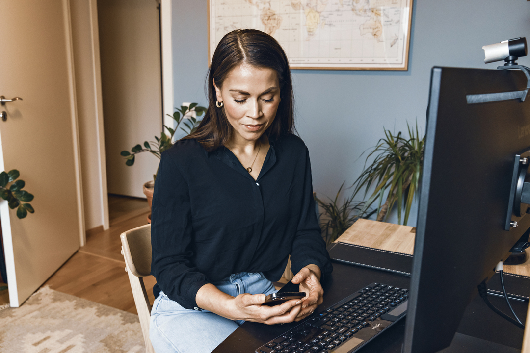Dame som sitter ved en PC. I hånden holder hun en mobil som hun ser ned på. Bilde.