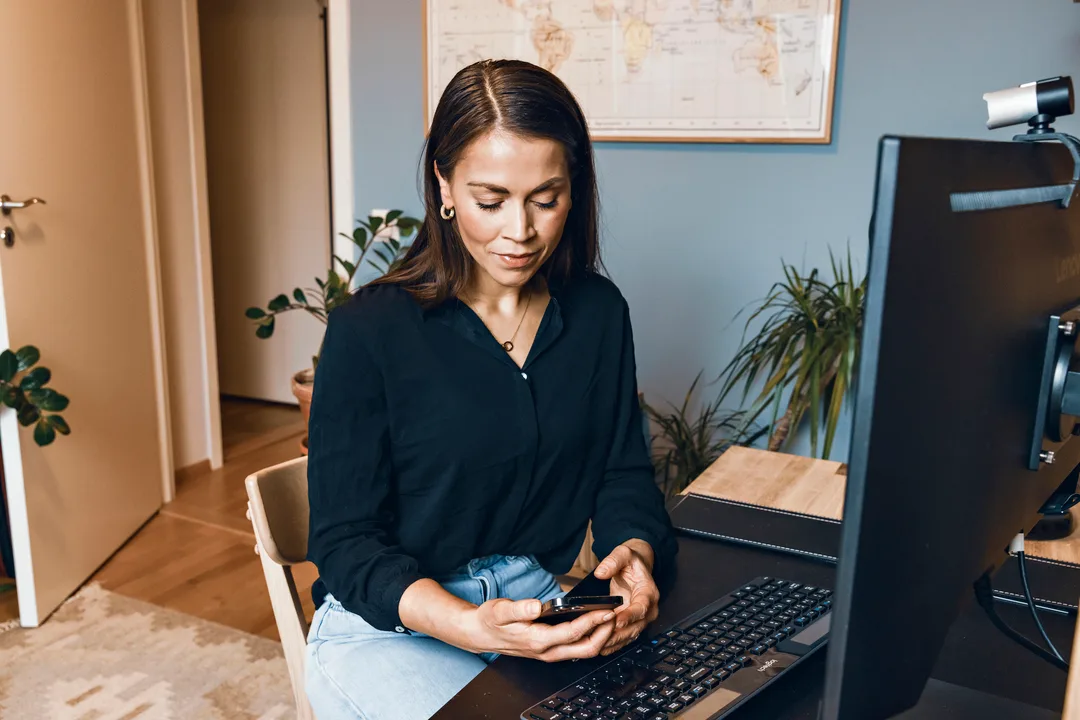Dame ved PC Dame som sitter ved en PC. I hånden holder hun en mobil som hun ser ned på. Bilde.