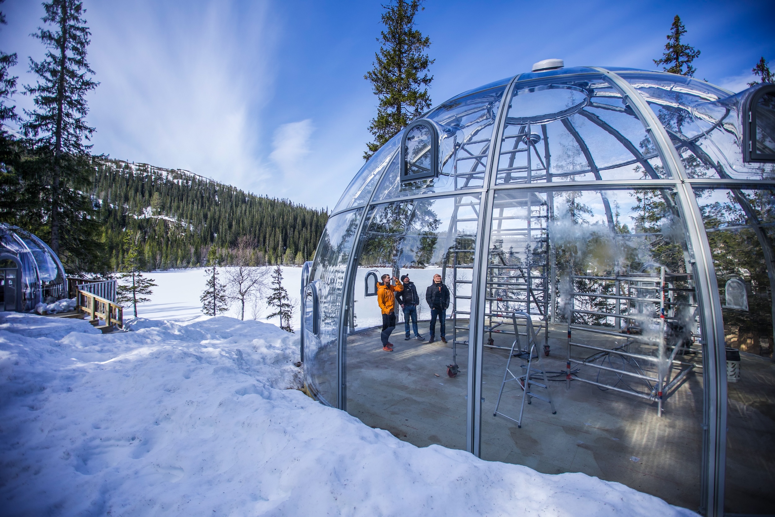 Tre menn står inne i en toetasjes gapahuk plassert midt i naturen med fjell, skog og snø på alle kanter
