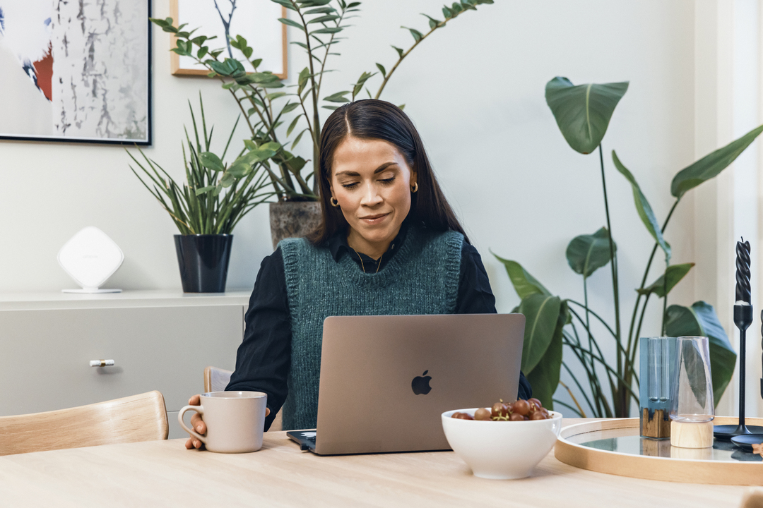 Same som sitter ved et bord. Hun ser på en laptop foran seg, og i hånden holder hun en kaffekopp. Bilde.