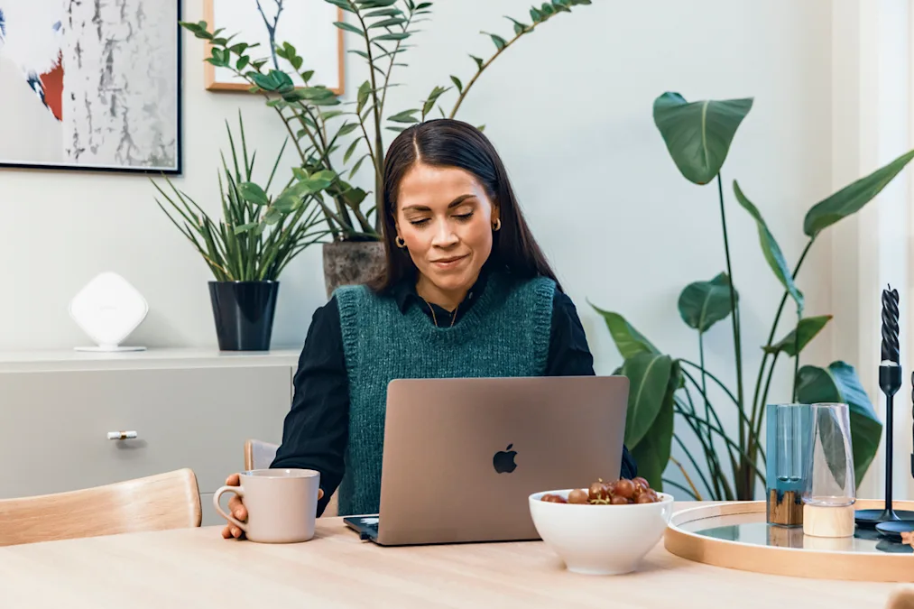 Dame som jobber på hjemmekontor Same som sitter ved et bord. Hun ser på en laptop foran seg, og i hånden holder hun en kaffekopp. Bilde.