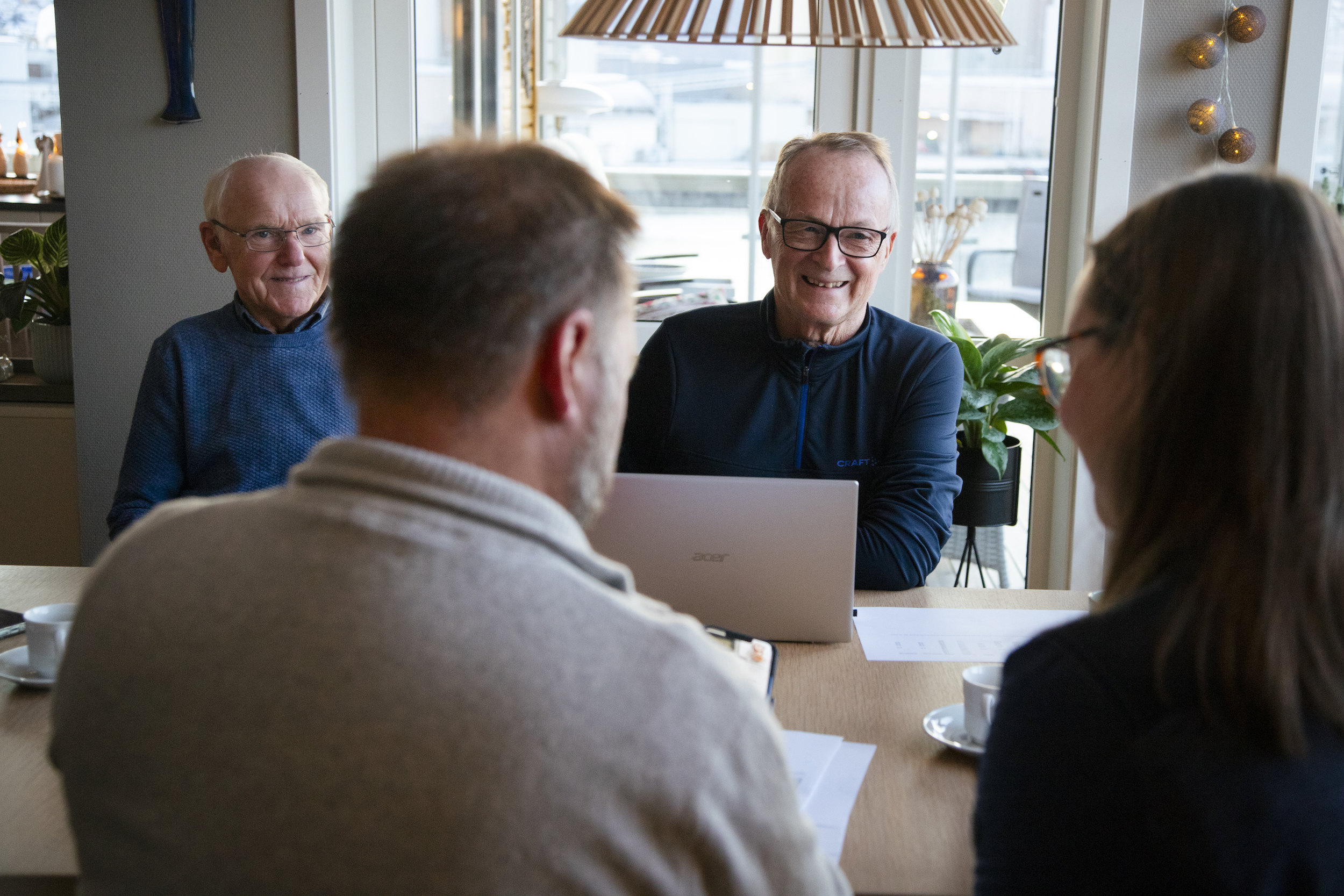 To smilende menn i samtale med en mann og en dame som sitter med ryggen til. 