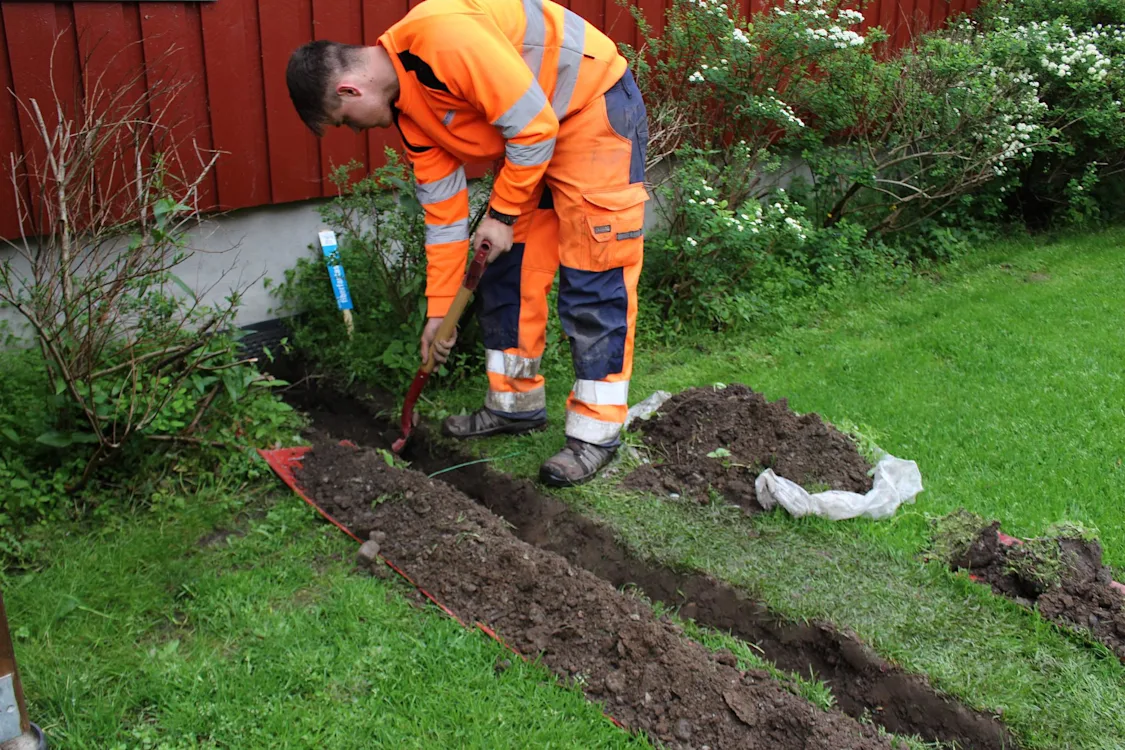 Kunden graver selv kabelgrøft for fiberkabel Mann i arbeidsklær som graver en kabelgrøft med en spade. Mannen står på en plen ved en husvegg og man ser at han har gravd en grøft i rett linje inn mot husveggen. Bilde.
