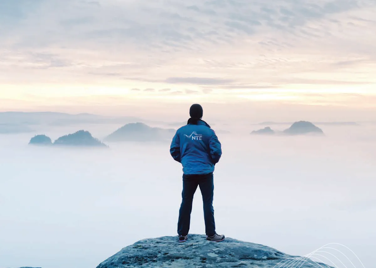 Bildet viser en mann stående på et berg og ser utover horisonten, mannen har en jakke med NTE-logo på ryggen Bildet viser en mann stående på et berg og ser utover horisonten, mannen har en jakke med NTE-logo på ryggen