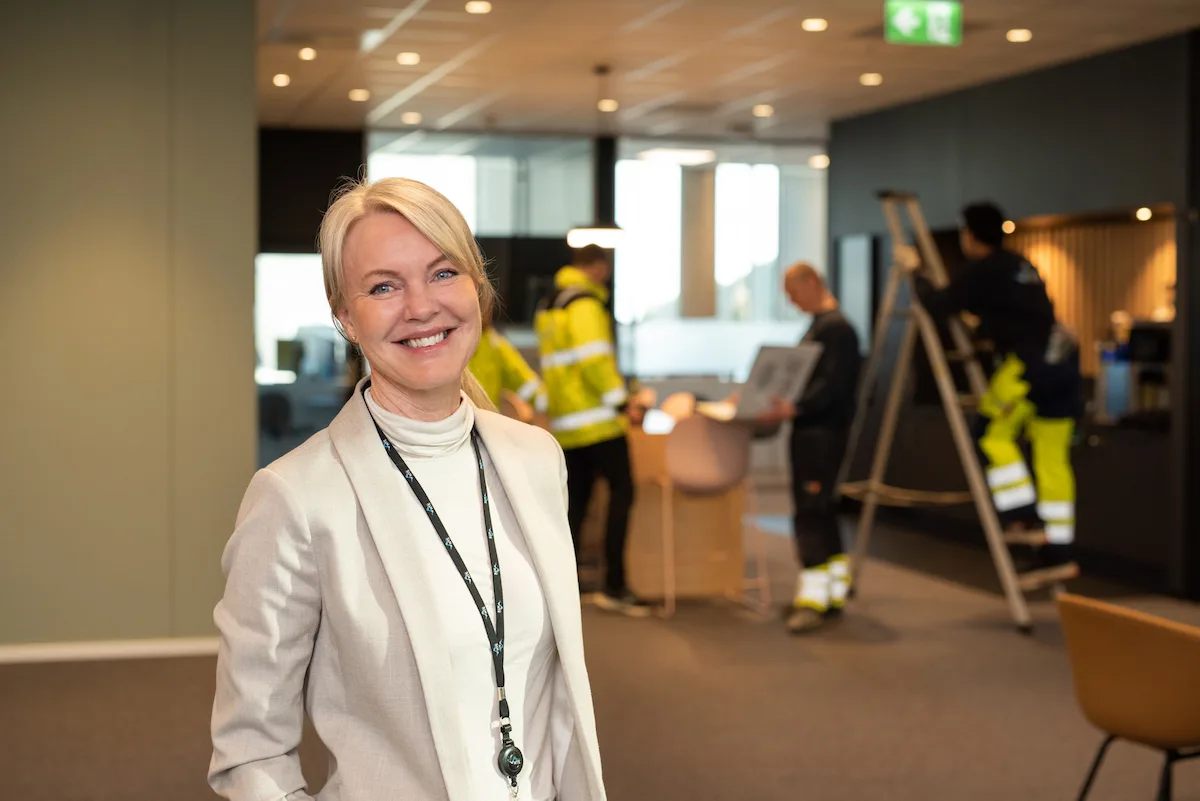 Camilla Berge Jacobsen 2 Smilende kvinne foran folk i arbeid på en gardintrapp og rundt et bord.