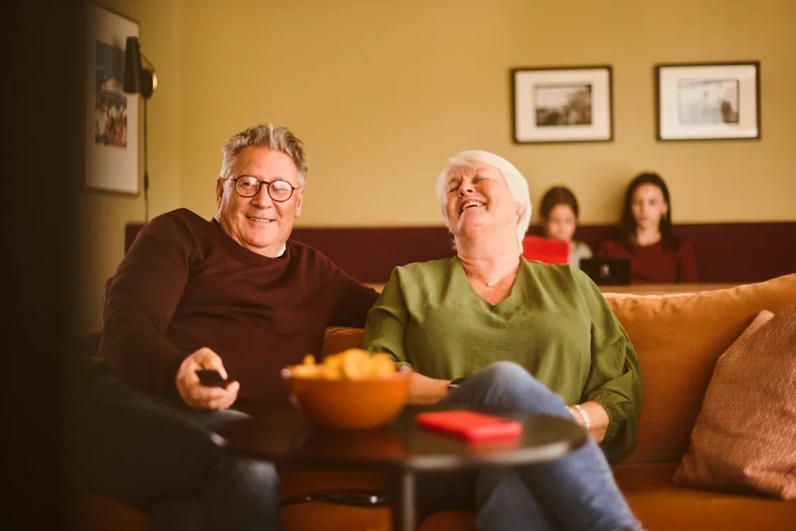 Besteforeldre ser tv – barnebarn ser på mobil. Foto: Andreas Kleiberg, Altibox Bilde av smilende besteforeldre som ser på tv. Barnebarn sitter i bakgrunnen og ser på egen skjerm.