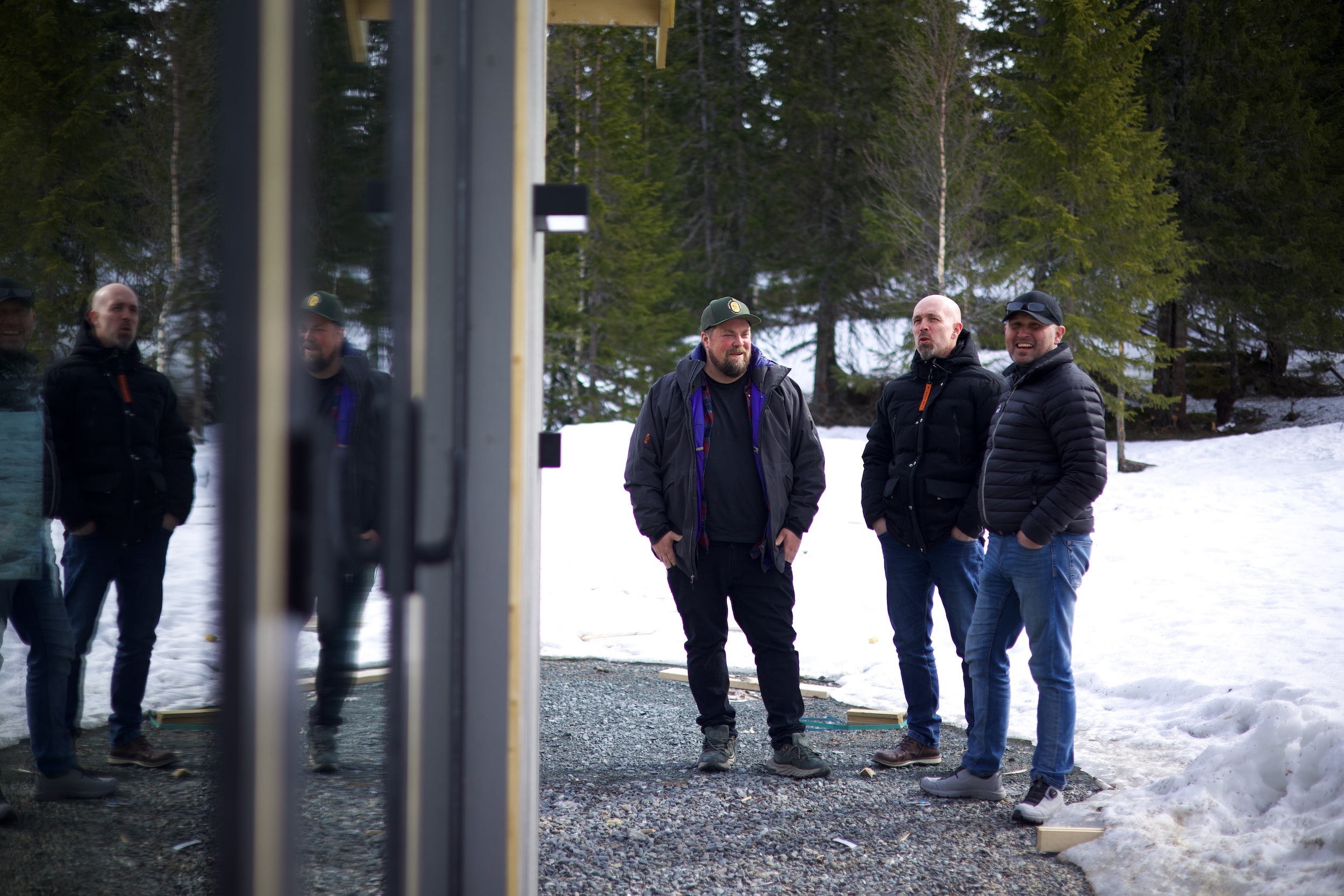 Tre menn står utenfor en hytte. Snø og skog i bakgrunnen. 