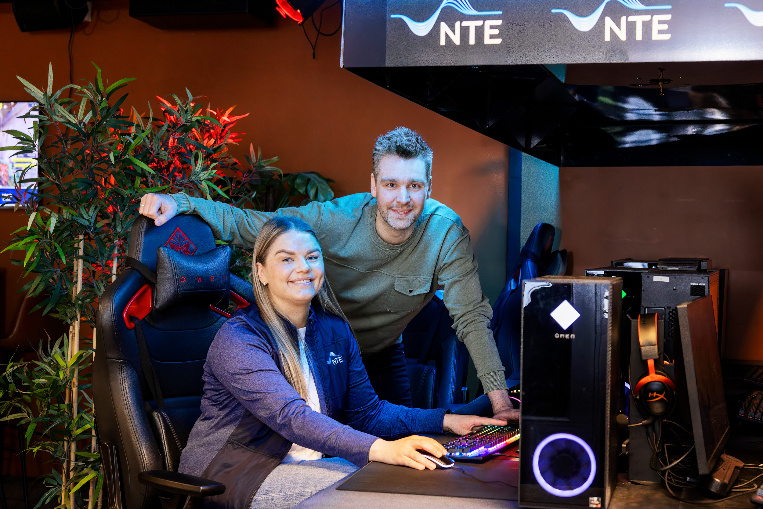 To smilende menn står på hver sin side av en blid dame. I bakgrunnen ser du ungdommer som sitter og gamer. 