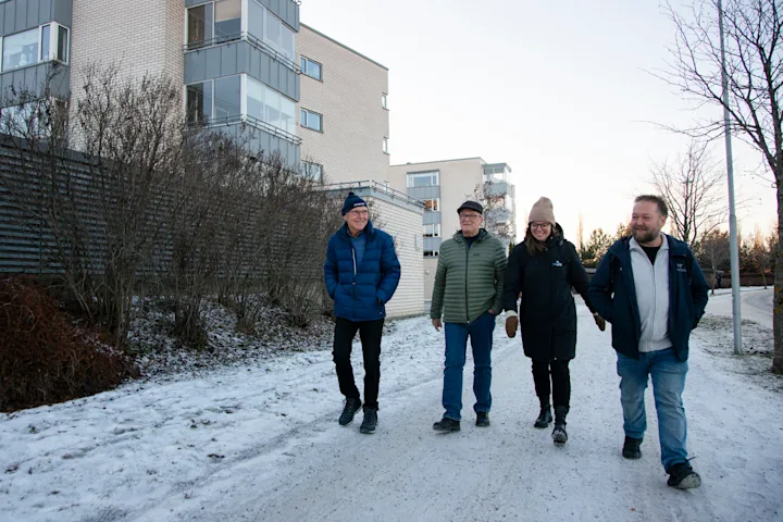 Tre menn og en dame, alle vinterkledde, går på fortauet utenfor et borettslag med mursteinsblokker. Tre menn og en dame, alle vinterkledde, går på fortauet utenfor et borettslag med mursteinsblokker.