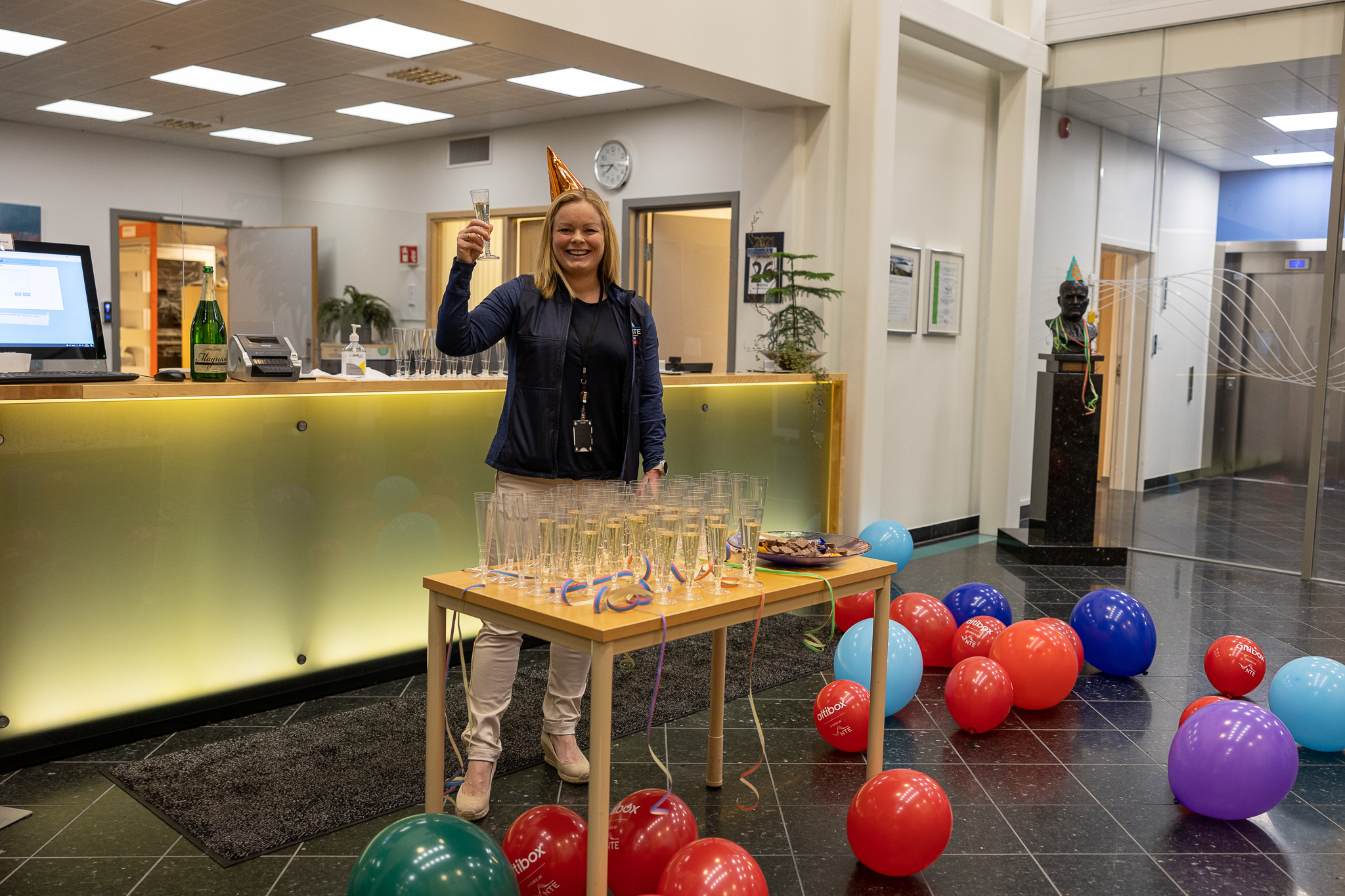 Kvinne med partyhatt står bak et bord med glass med brus og sjokolade. På gulvet ligger mange oppblåste ballonger. 