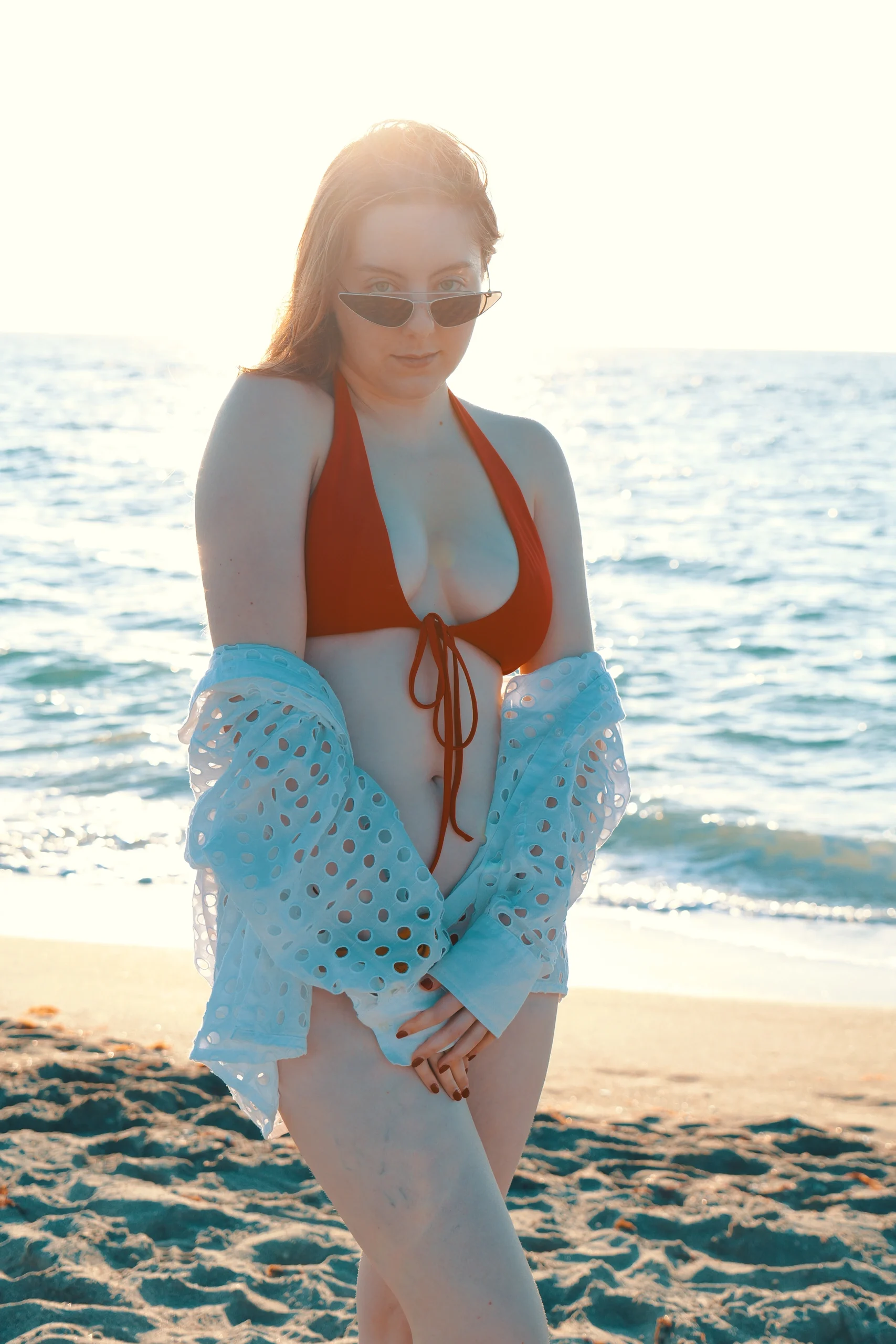 Model in orange halter swimsuit with blue perforated coverup and sunglasses on beach