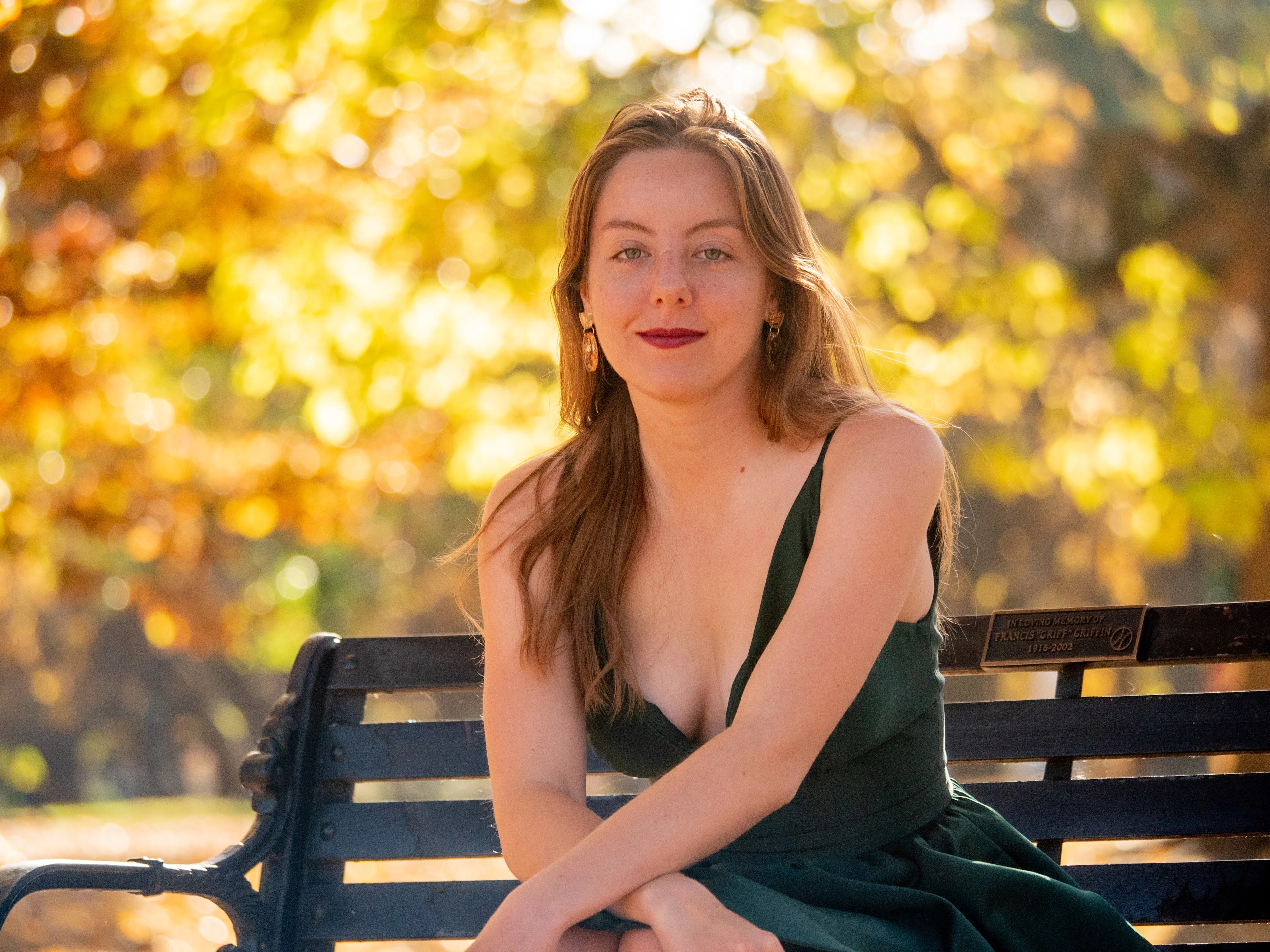 Model on park bench in emerald dress, hand in hair, warm golden hour light, autumn foliage bokeh