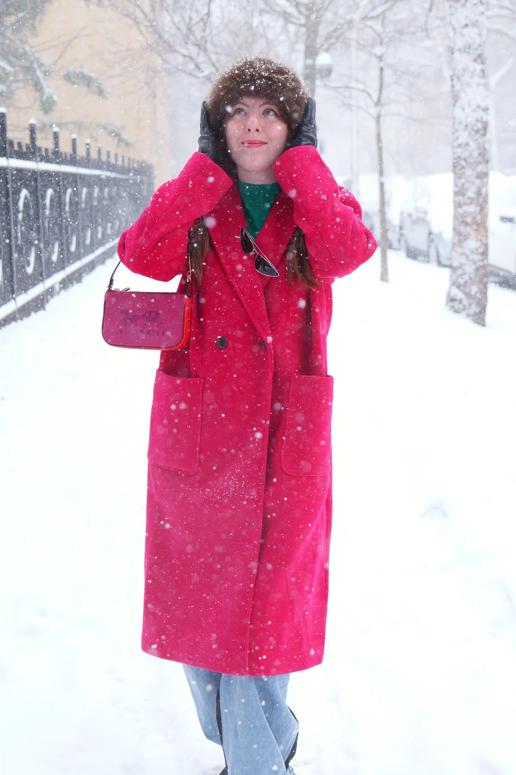 Model in pink winter coat adjusting fur-trimmed hood with upward gaze during active snowfall