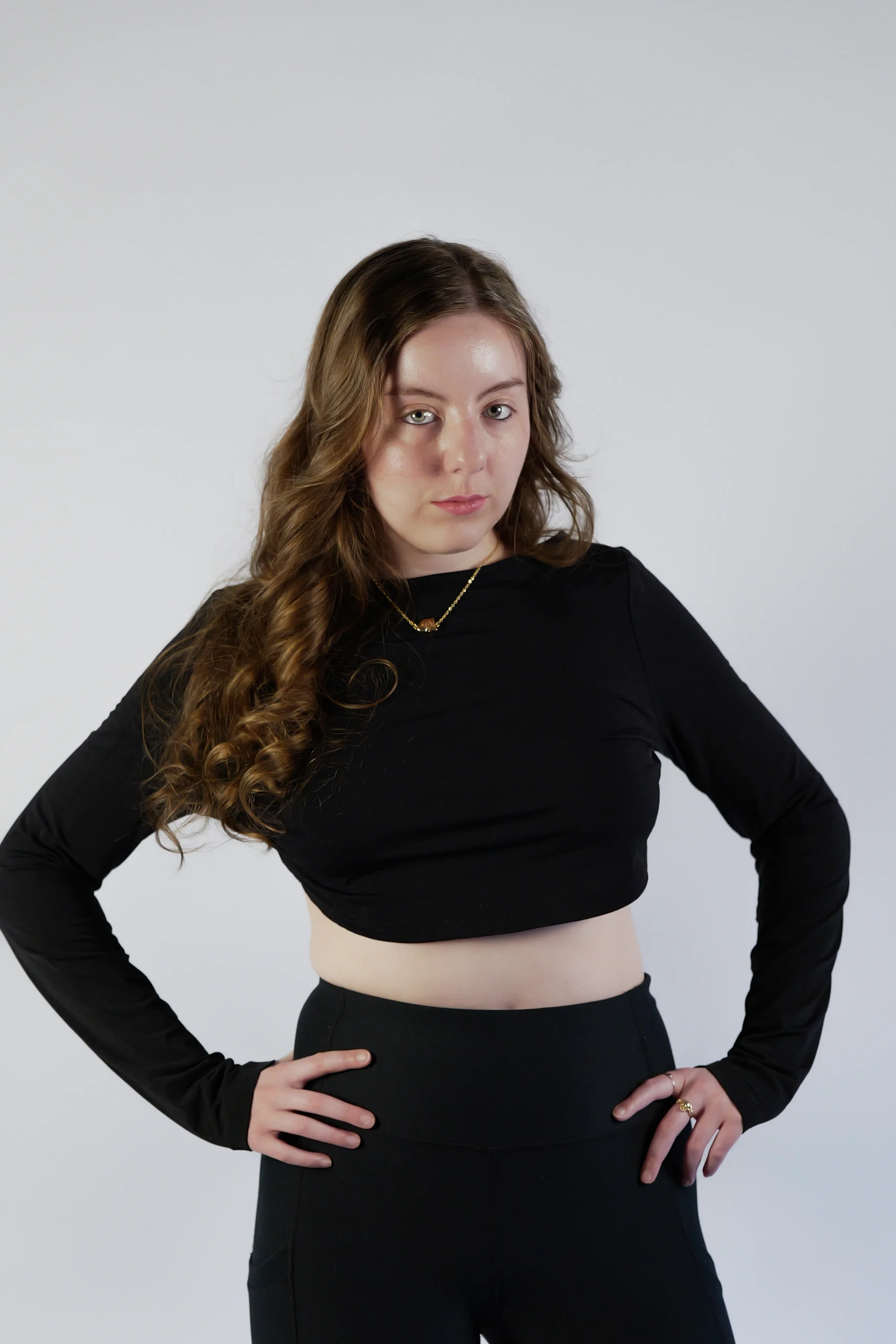 Hands-on-hips pose in black outfit with gold necklace, wavy hair, white studio backdrop