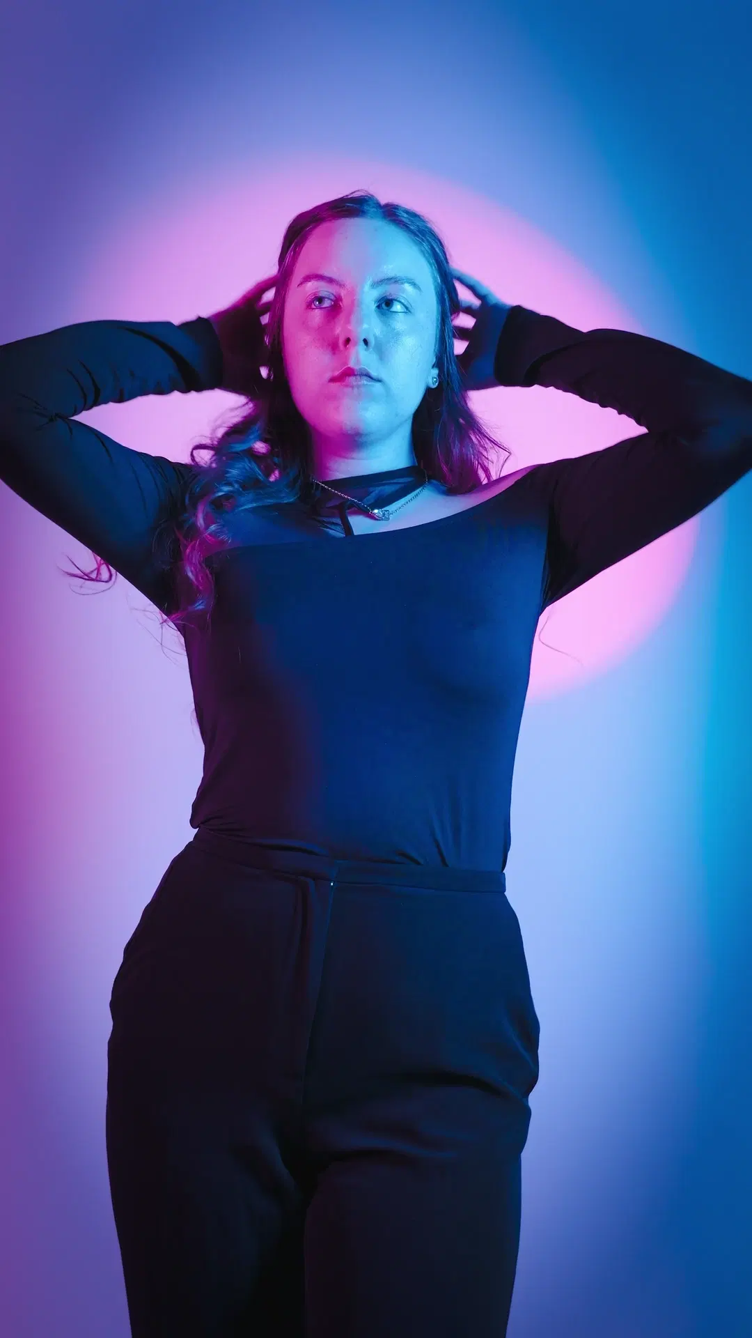 Studio portrait with magenta-cyan gel lighting, hands behind head pose, dark wardrobe