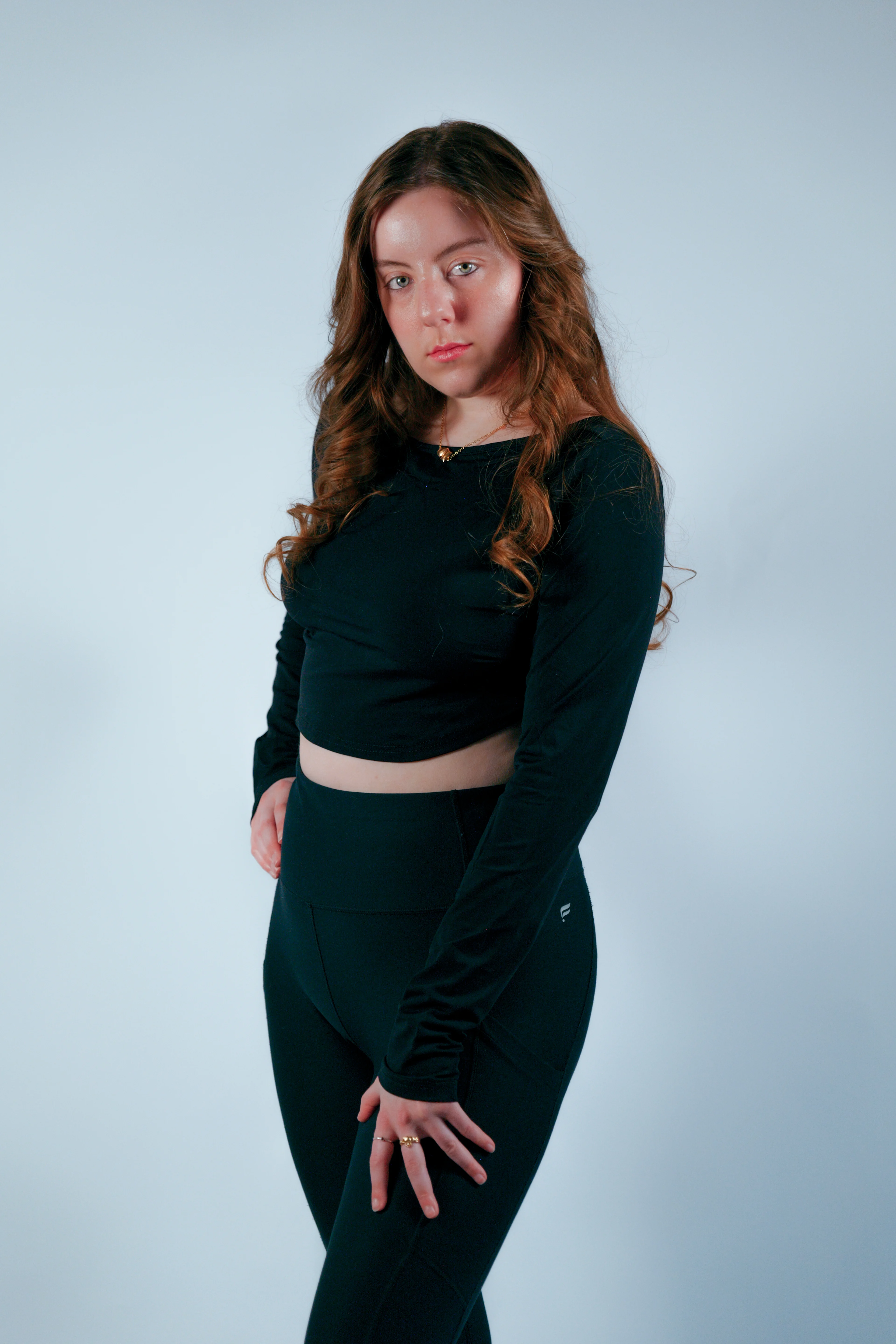 Three-quarter pose with wavy auburn hair, black crop top and leggings, soft blue studio backdrop