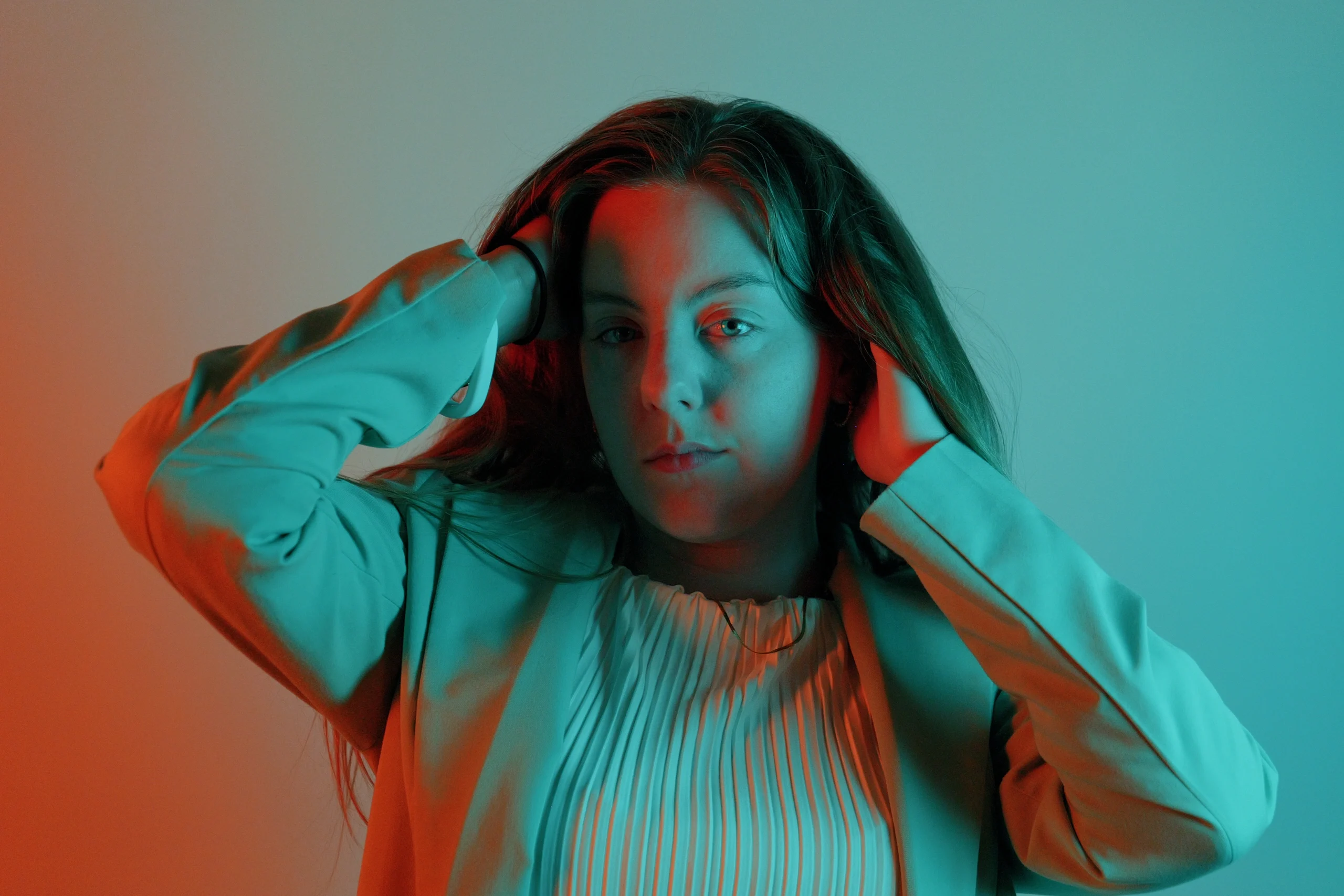 Studio portrait with cyan-orange gel lighting, hands raised to hair, white pleated top