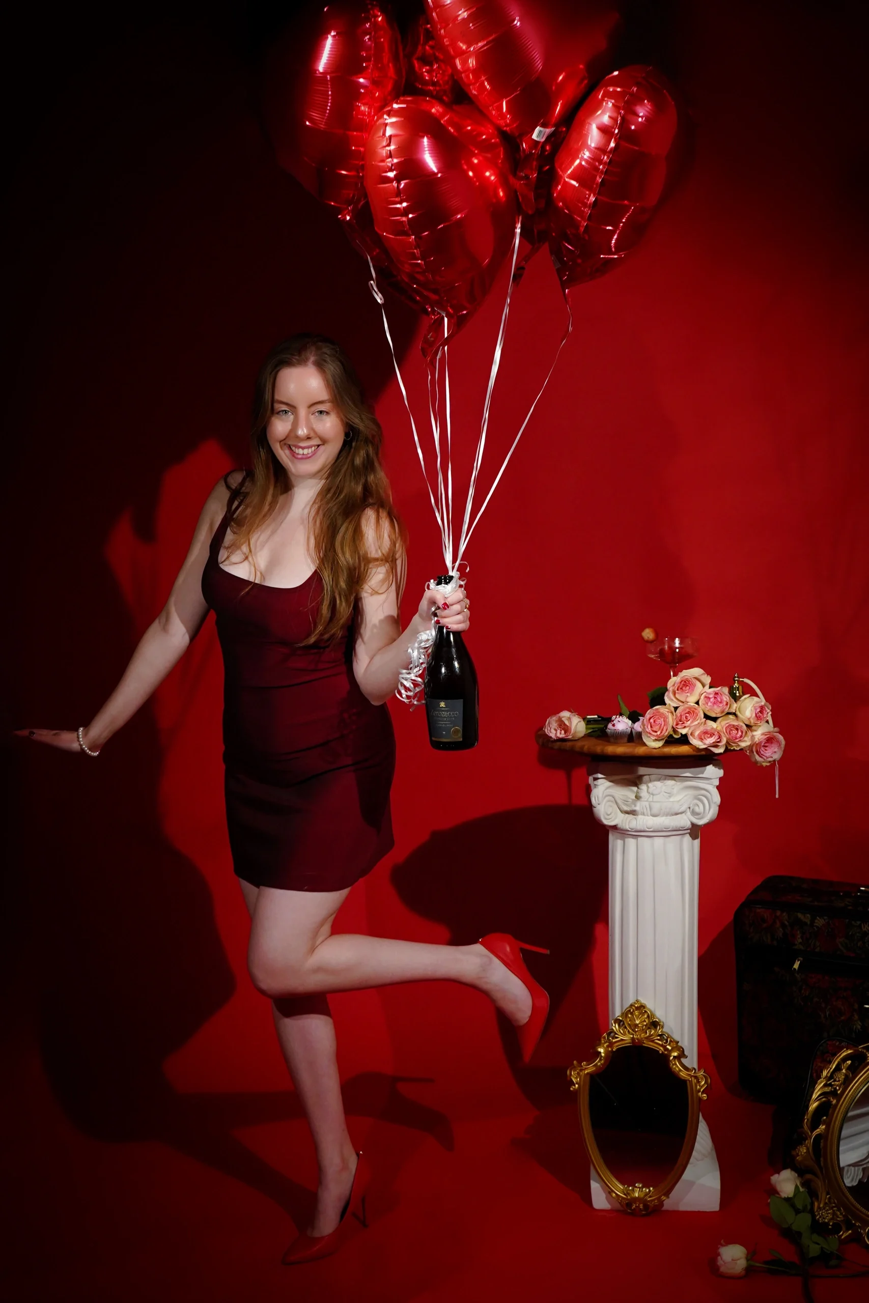 Dynamic pose in burgundy dress holding red heart balloons and champagne on red backdrop with roses