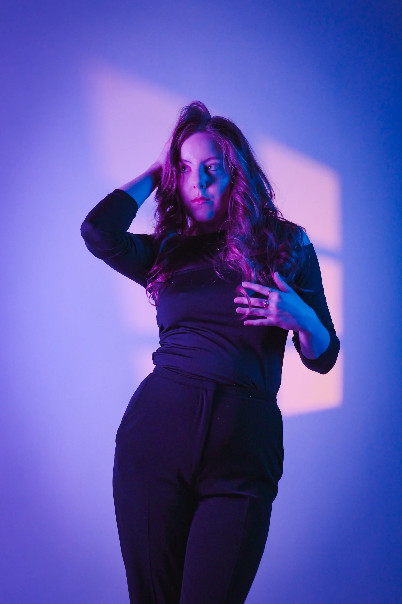 Editorial pose in black outfit, hand in hair, with magenta-cyan gradient gel lighting