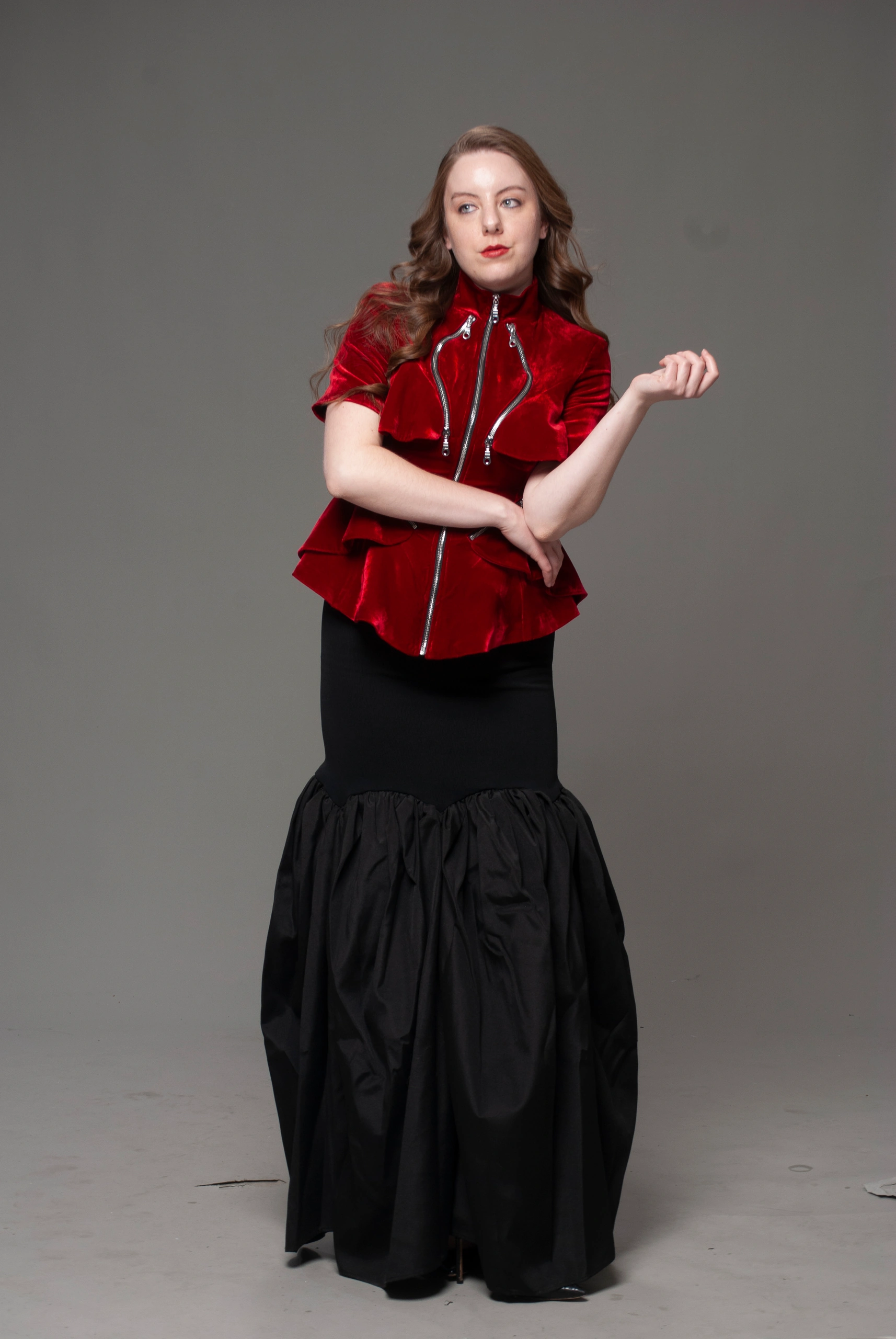 Full-length studio portrait in red velvet zip-up jacket and black gathered-skirt gown on gray backdrop