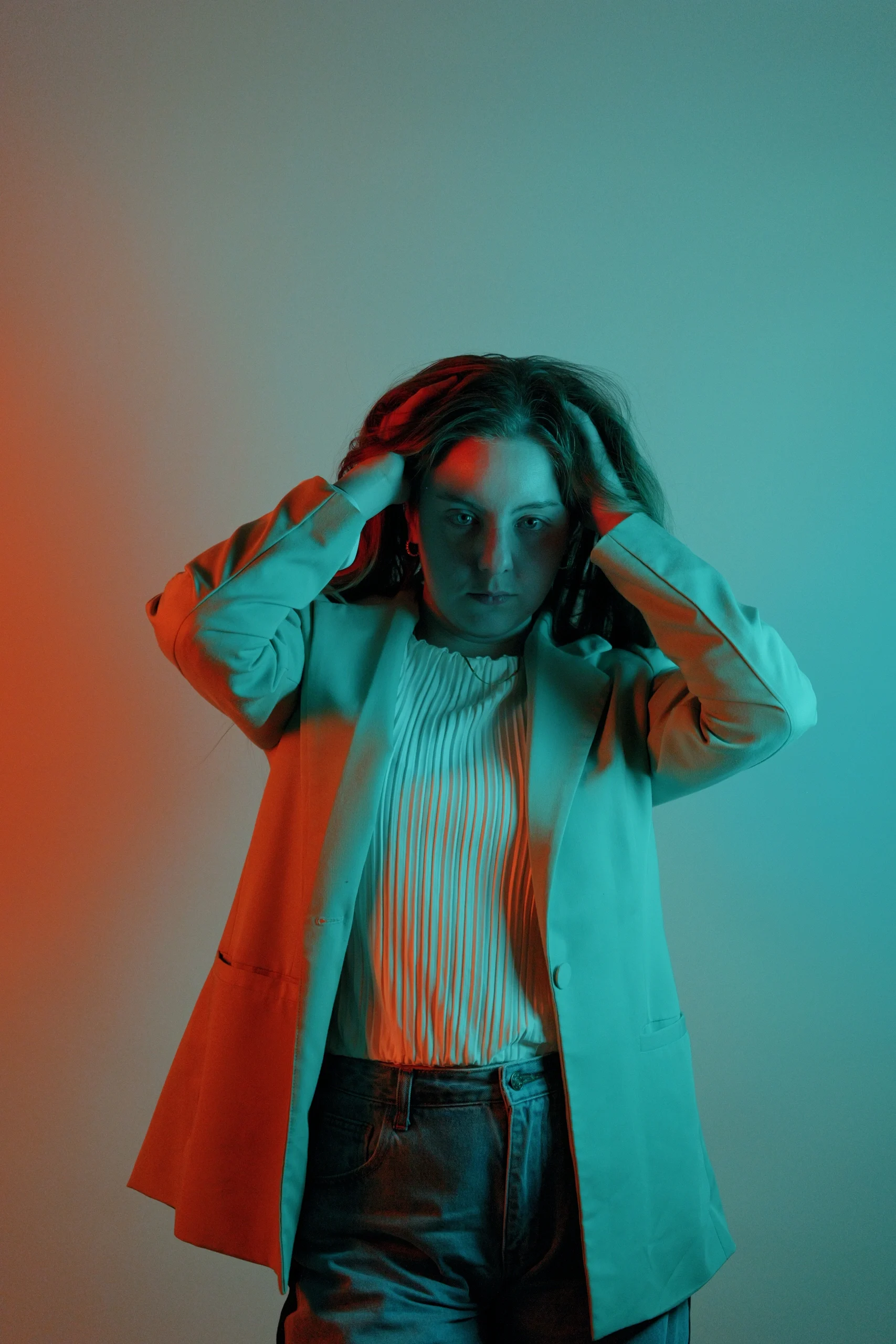 Studio portrait with orange and cyan split lighting, white blazer, pleated top, hands in hair