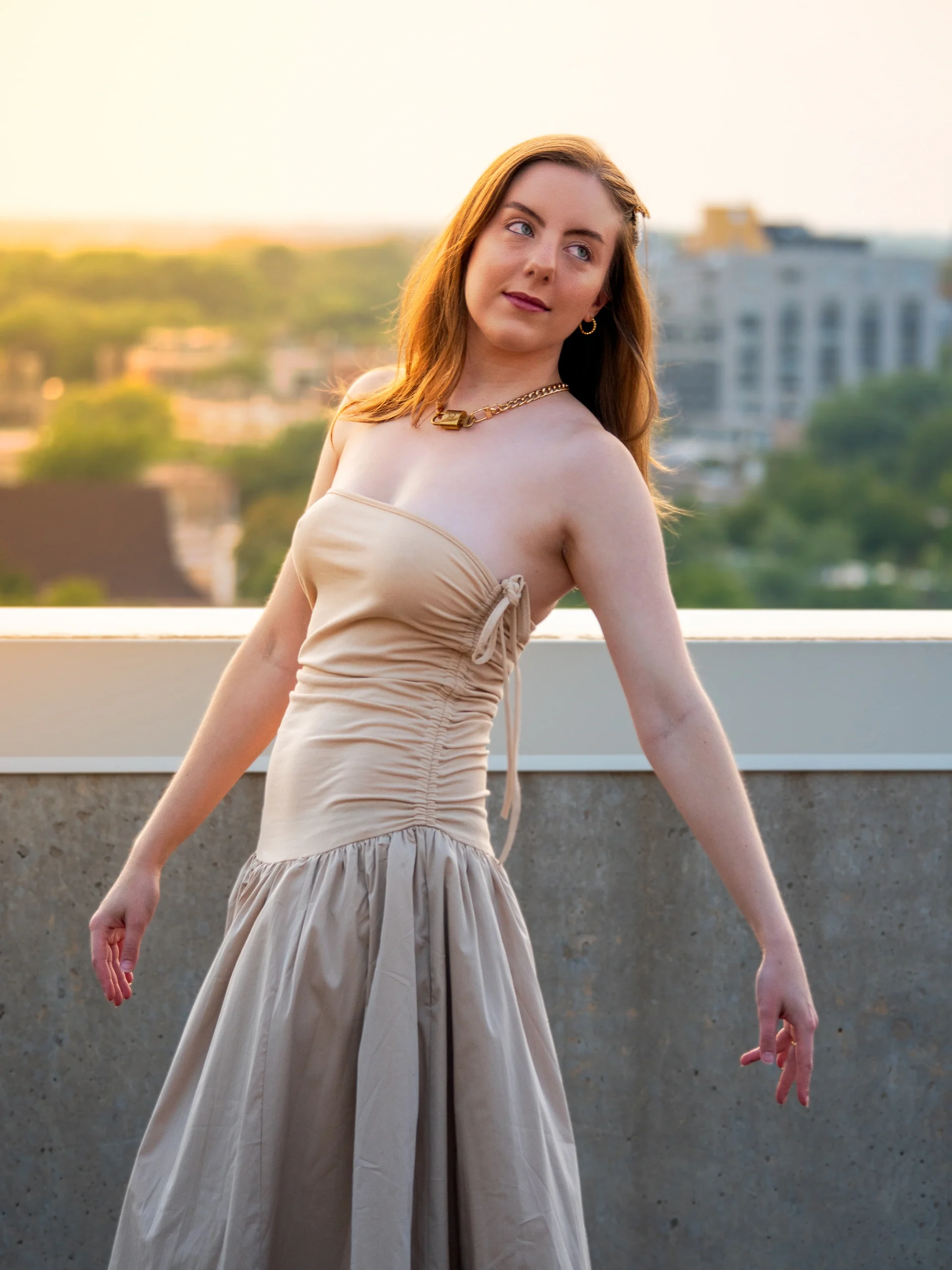 Beige evening gown on rooftop at sunset with over-shoulder pose, warm golden light, and city view
