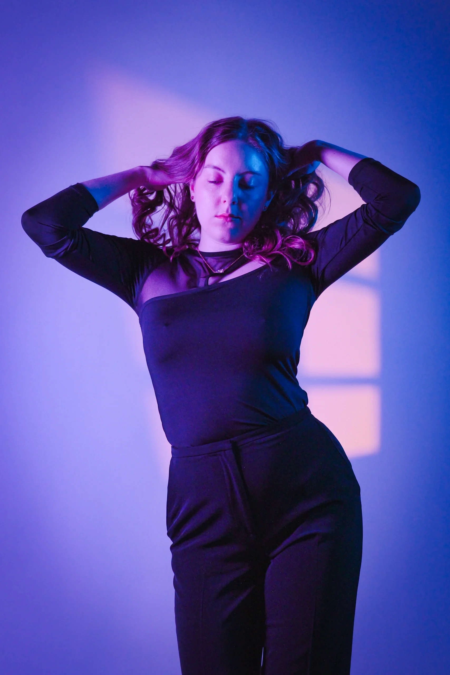 Editorial pose in black outfit with hands in hair under magenta-cyan studio lighting
