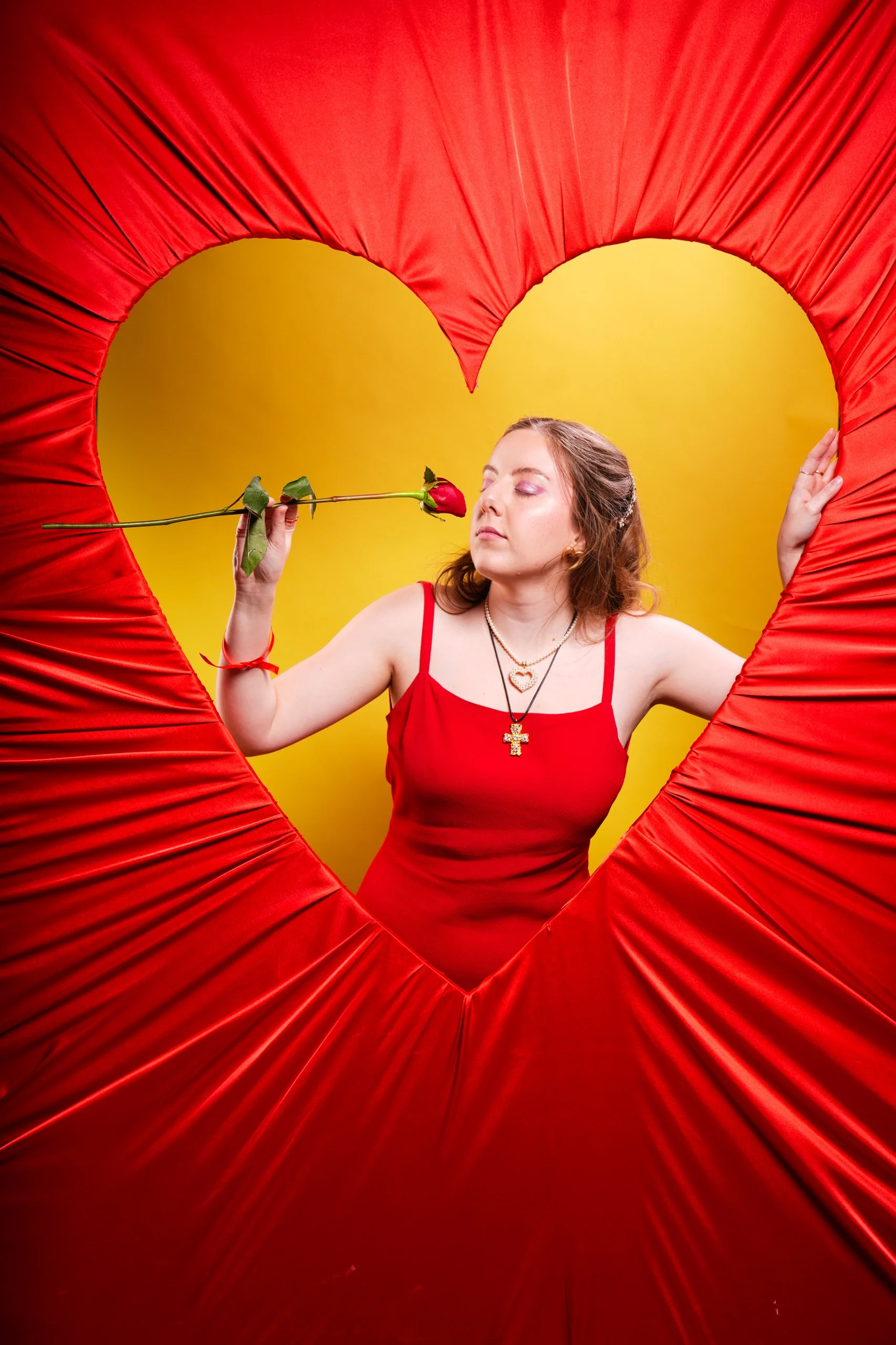 Model centered in gold heart frame, arms spread, holding red rose, red pleated backdrop