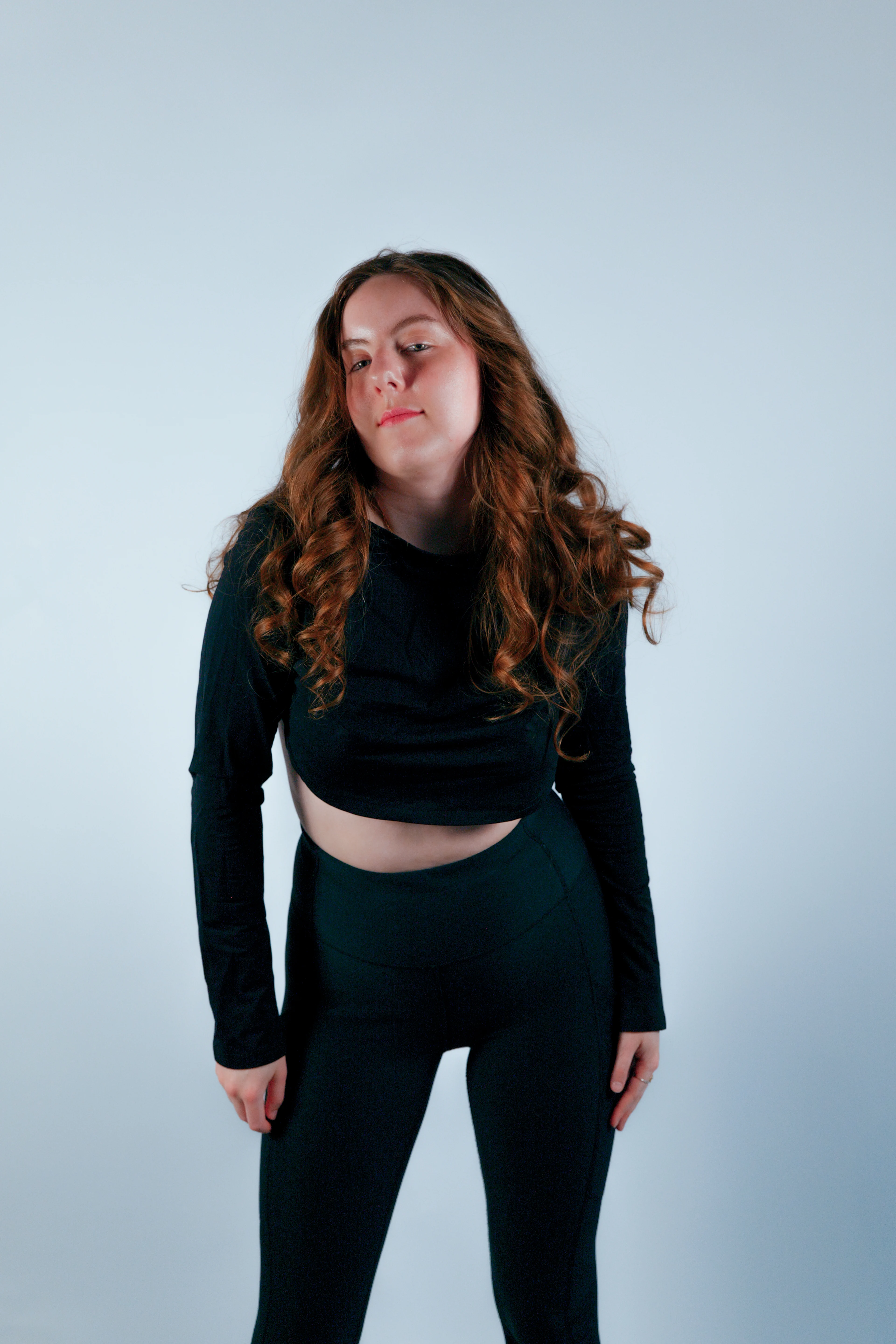 Black long-sleeve crop top and pants, flowing auburn curls, head tilted back on pale blue backdrop