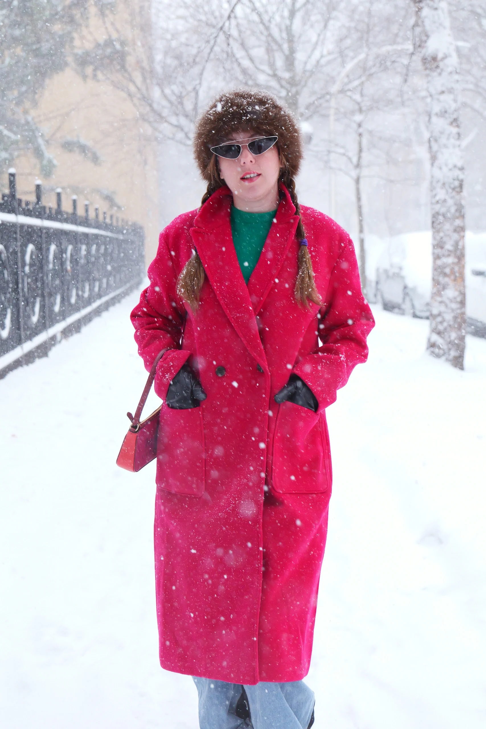 Model in red double-breasted coat, brown fur hat, and cat-eye sunglasses during urban snowstorm