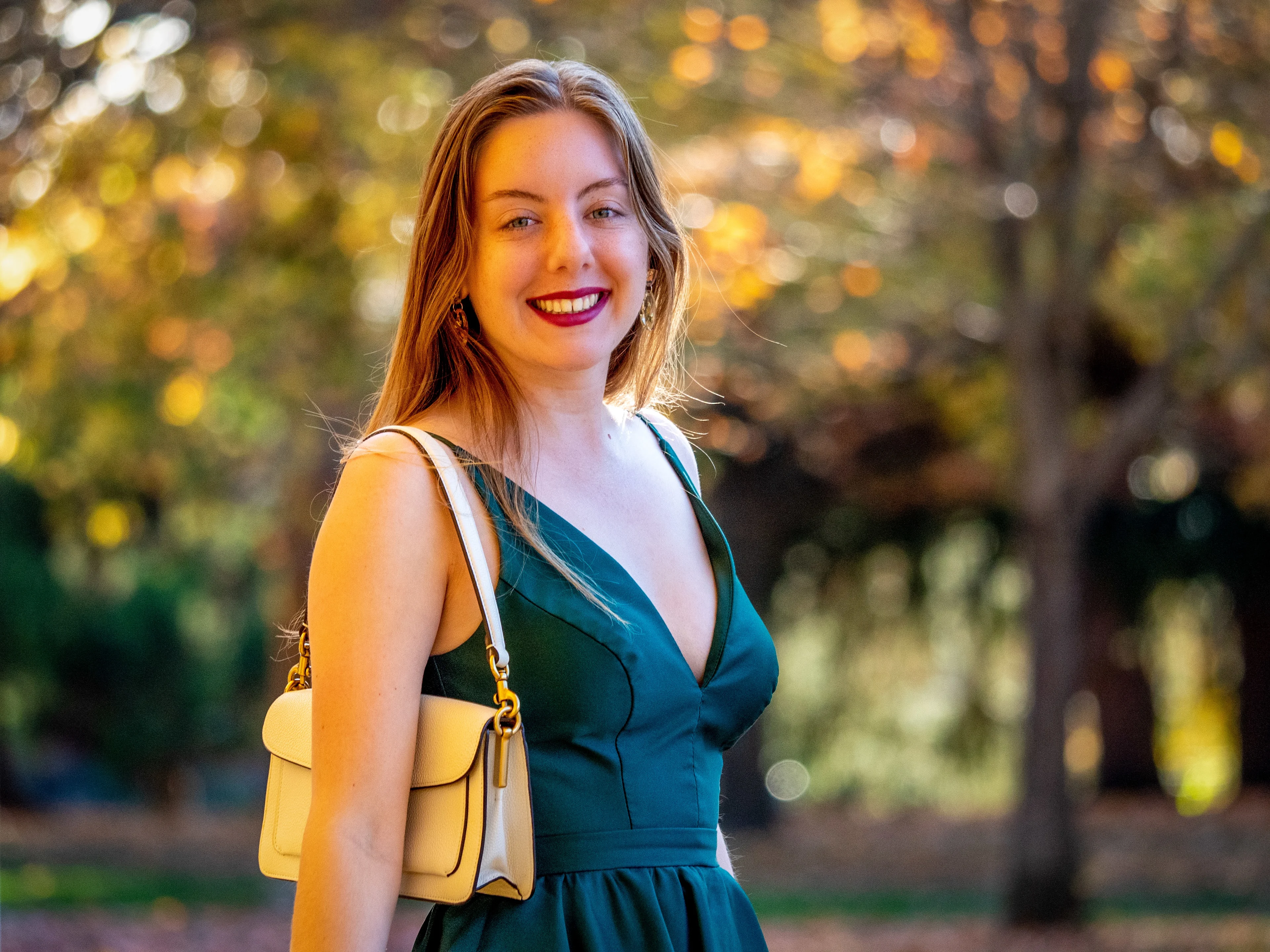 Model in teal dress with yellow shoulder bag, warm golden hour lighting, blurred autumn foliage