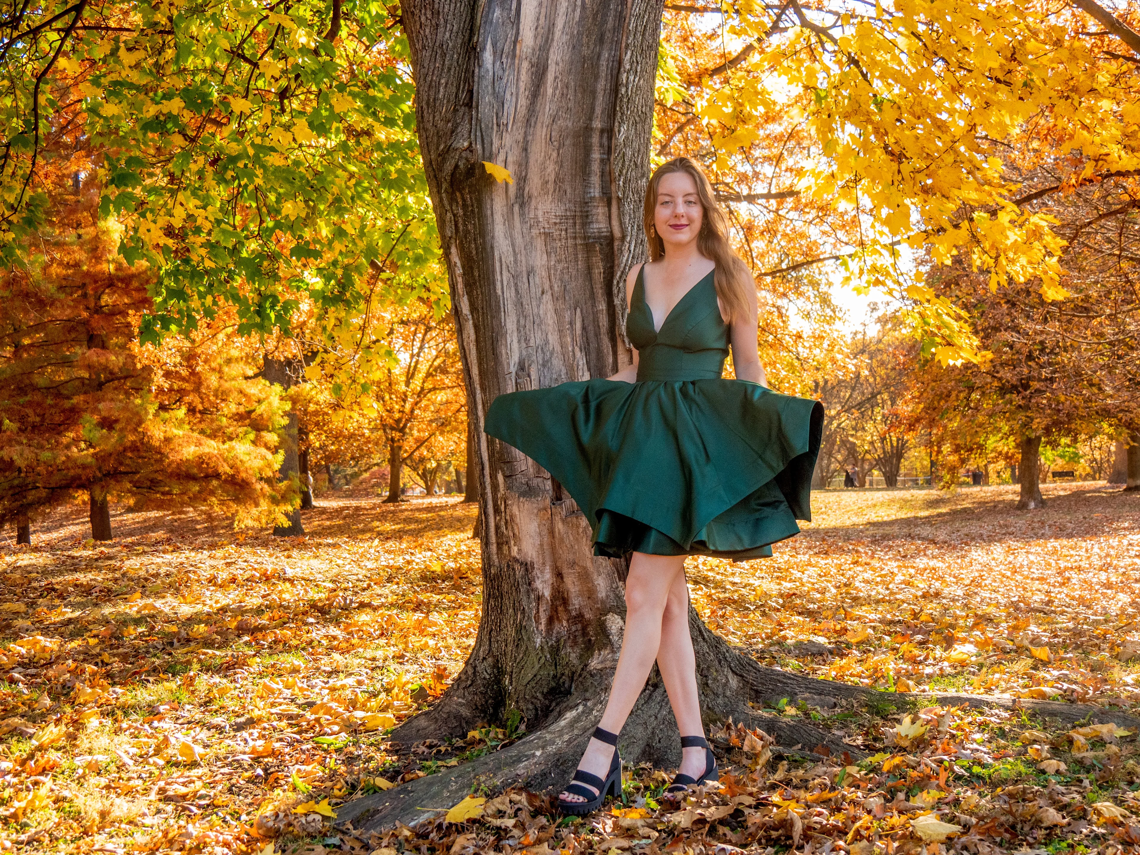 Model in flowing emerald V-neck dress posed at tree trunk with golden fall foliage and warm lighting