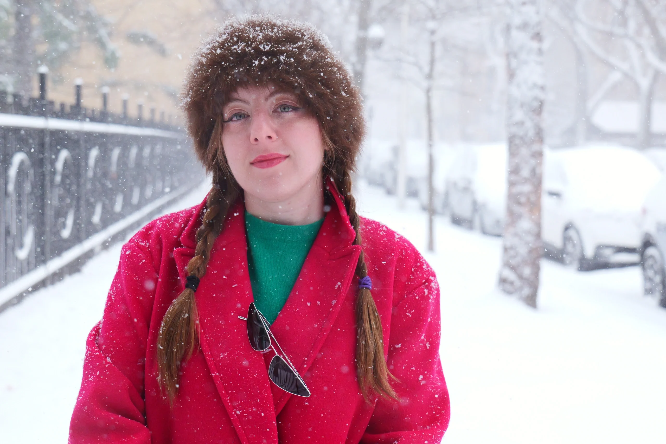 Portrait in red coat and fur hat with braided hair during active snowfall on urban street