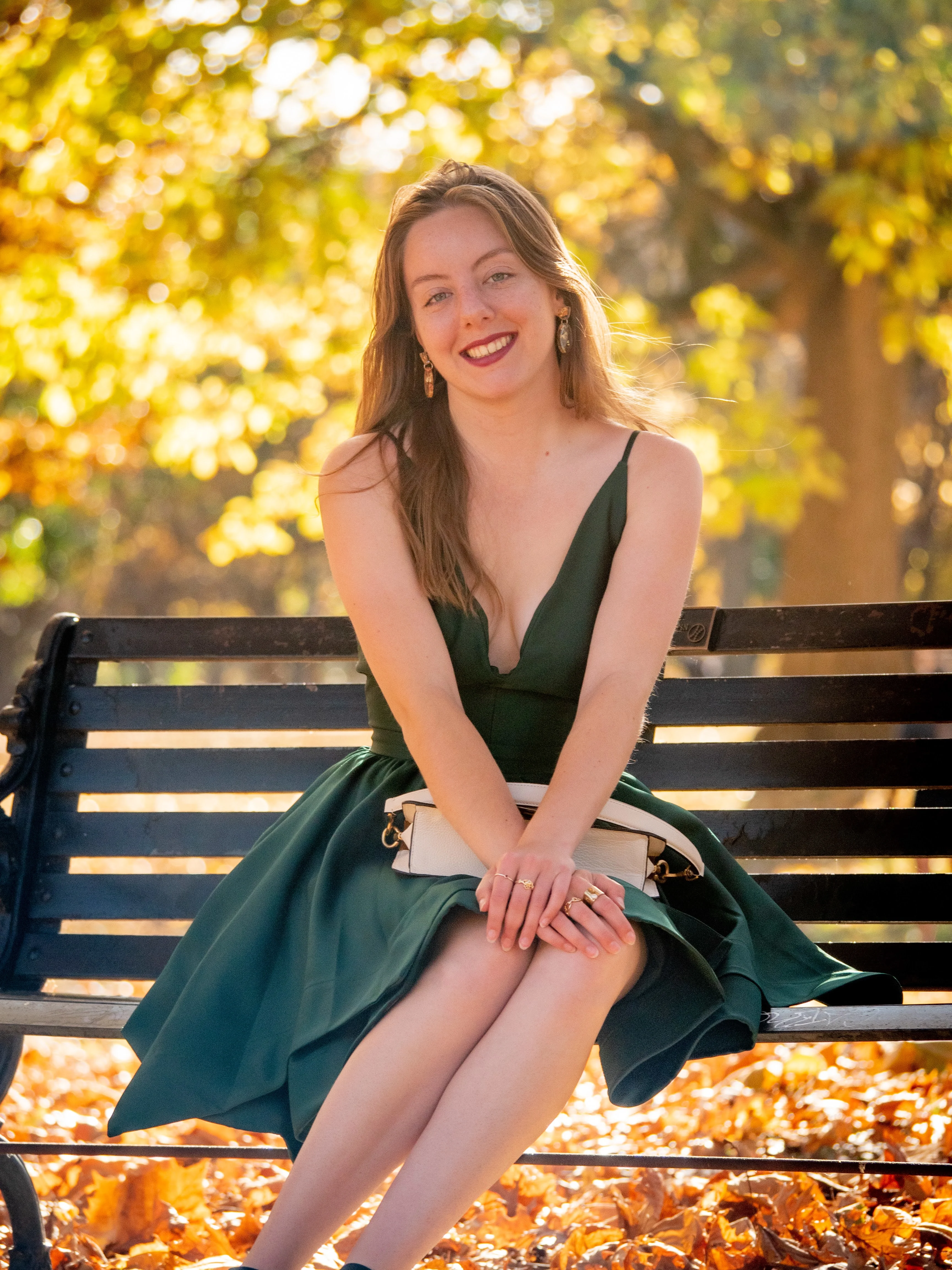 Model in emerald dress seated on park bench with warm golden light and autumn foliage bokeh