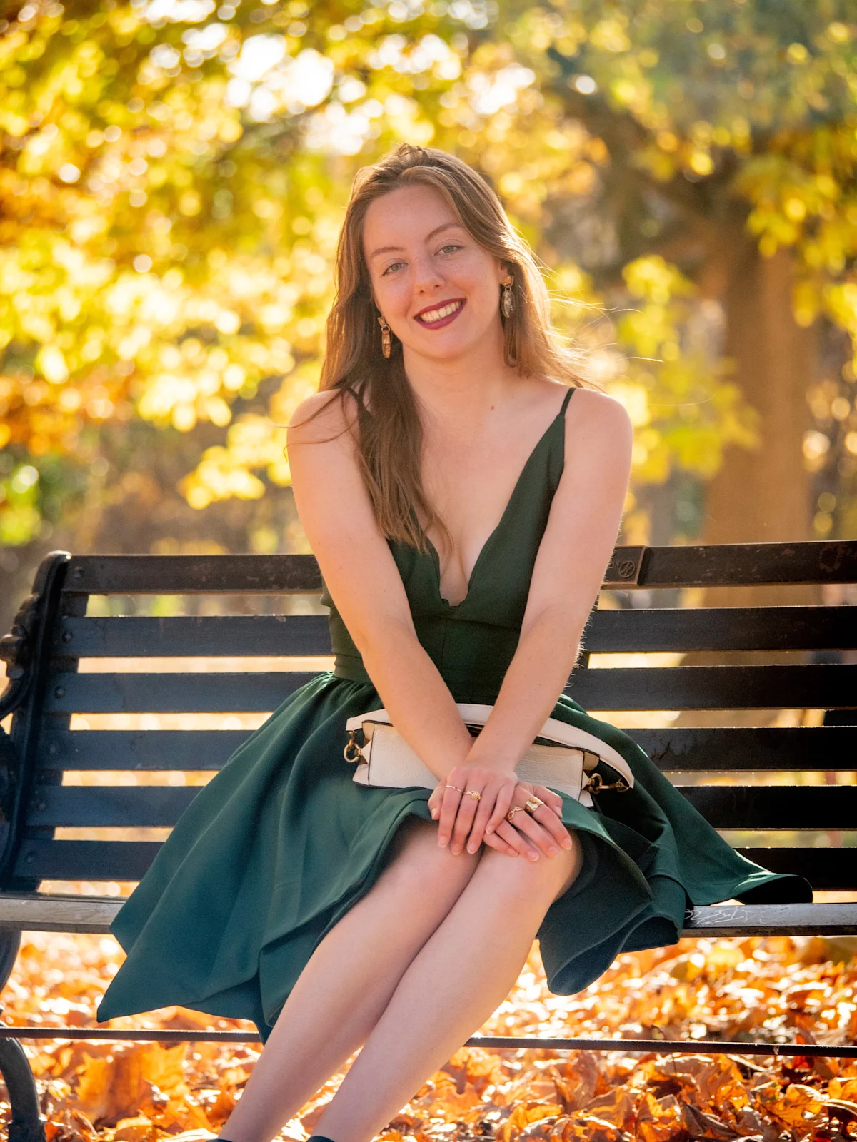 Model in emerald dress seated on park bench with warm golden light and autumn foliage bokeh