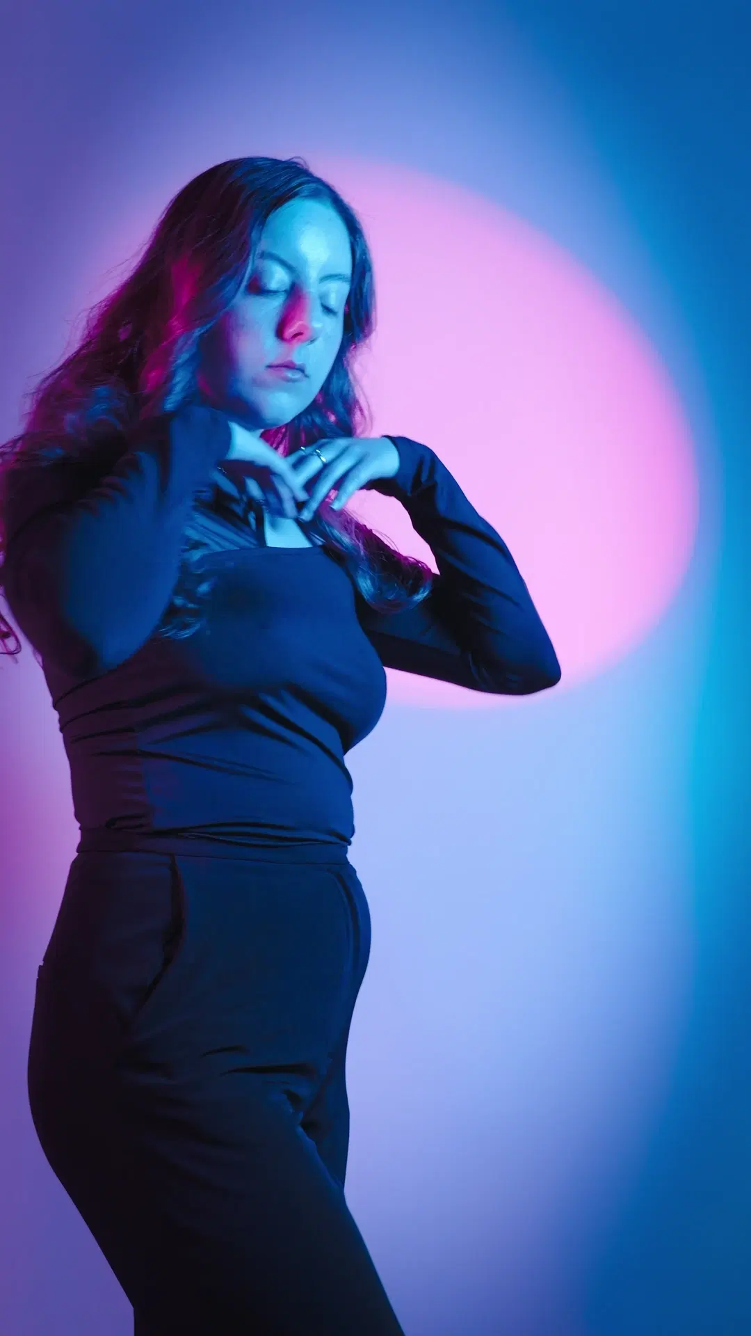 Eyes closed, hands at chest in black outfit against magenta-pink halo and cyan gradient