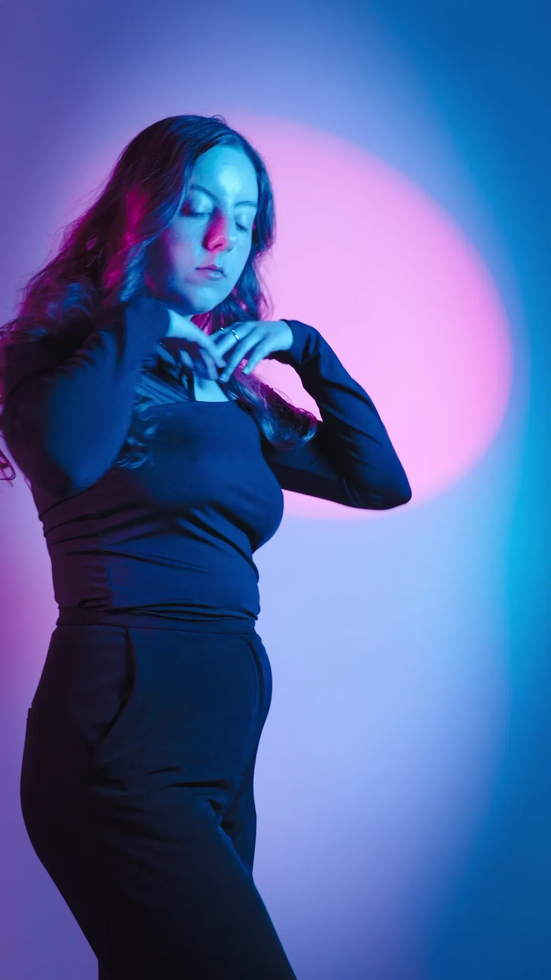 Eyes closed, hands at chest in black outfit against magenta-pink halo and cyan gradient